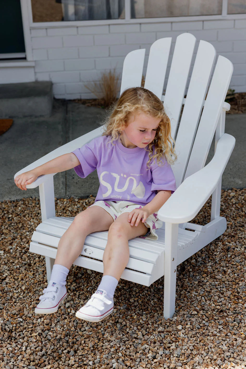 A young child with curly blonde hair sits thoughtfully on a white Adirondack chair outdoors, wearing an oversized fit SUNDAY SIBLINGS Il Sole Tee in purple and cream, shorts, and sneakers, while gazing at the gravel around them.