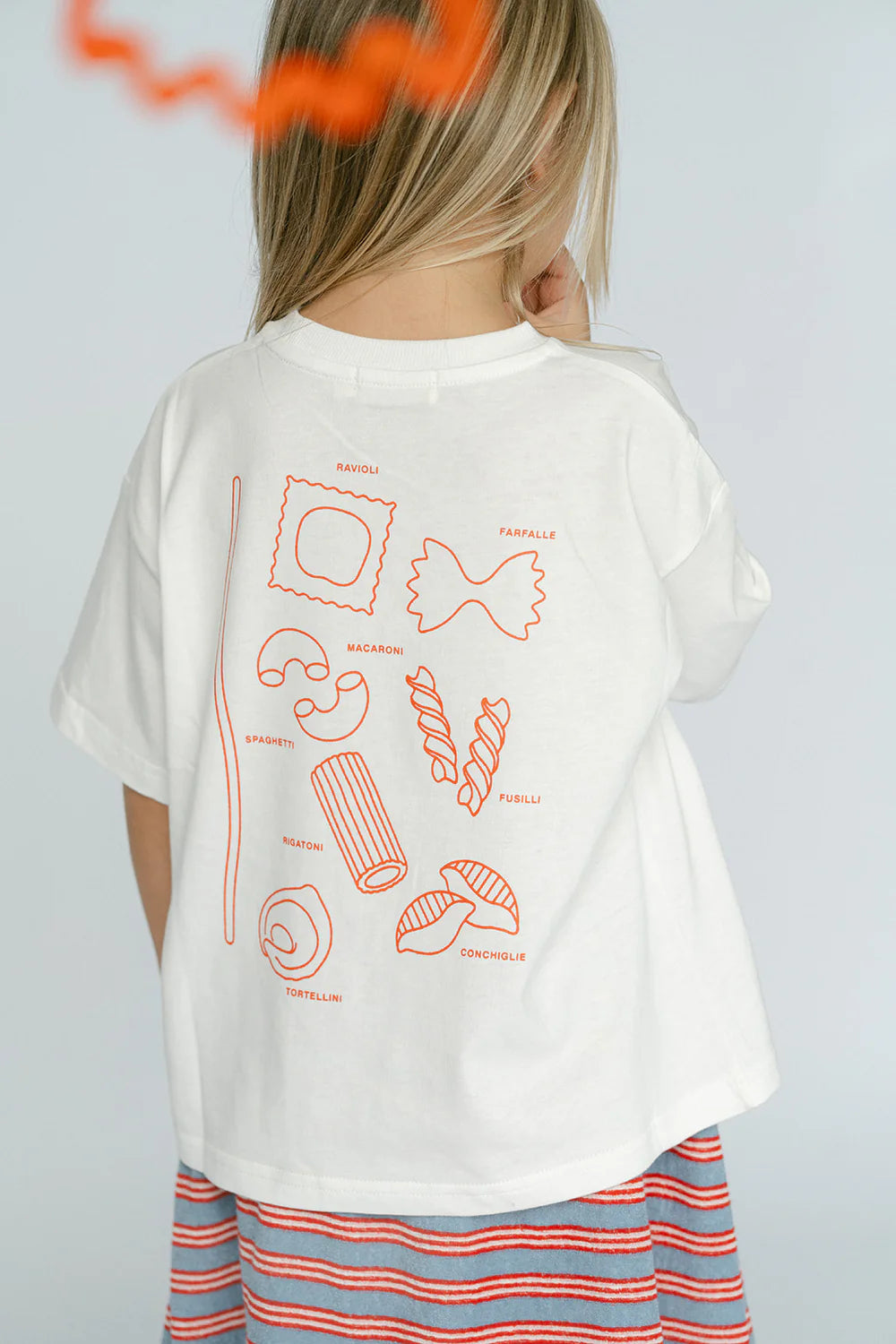 A child with light-colored hair is wearing an oversized fit white Pasta Tee by LENN LABEL, crafted from soft cotton jersey and decorated with orange illustrations and names of various pasta shapes on the back. The child has paired it with a striped skirt featuring red, blue, and white stripes. The background is plain and light-colored.