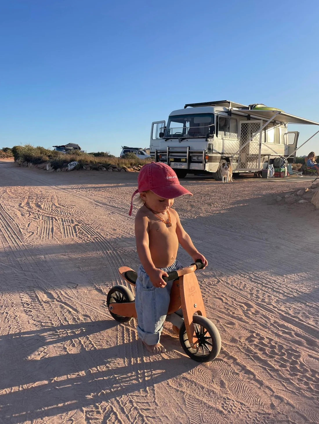 A child in blue pants and a ZIGGY ZAZA Roses Are Red cap rides a wooden balance bike on a sandy path. An RV is parked under the clear blue sky, surrounded by sparse vegetation and dirt paths, with an adjustable brass clasp glinting softly in the sunlight.