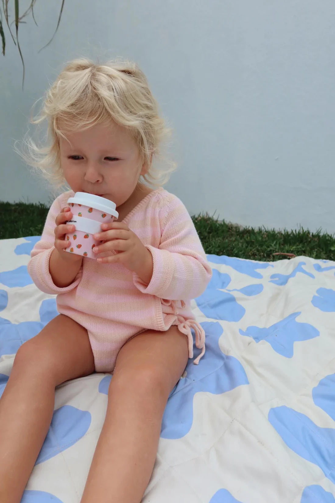 A toddler with blonde hair sits comfortably on a blue and white blanket outdoors, wearing the MILKY DESIGNS Essential Wrap Romper in Pink/Peach. The child sips from a white cup with a floral pattern, with a wall in the background and grass nearby.
