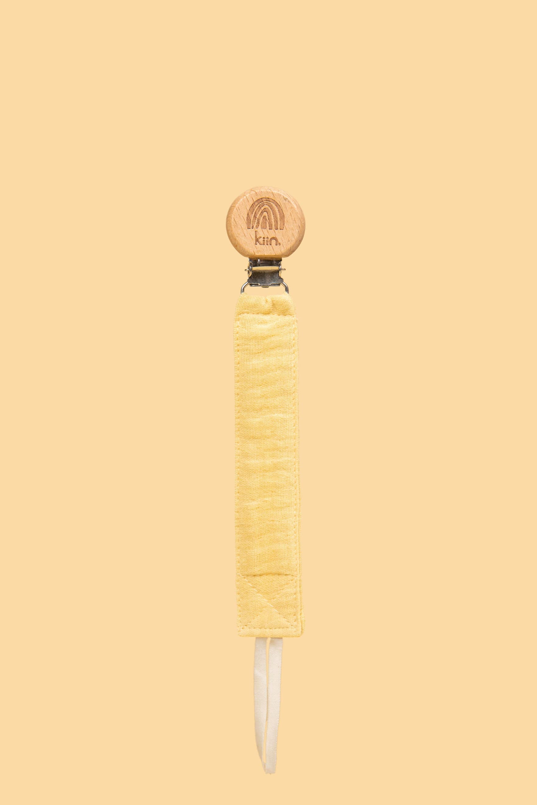 A wooden pacifier clip engraved with "Kiin" is attached to a Kiin Baby cotton dummy clip strap in yellow. The matching yellow background makes it a perfect baby shower gift for a gender-neutral nursery.