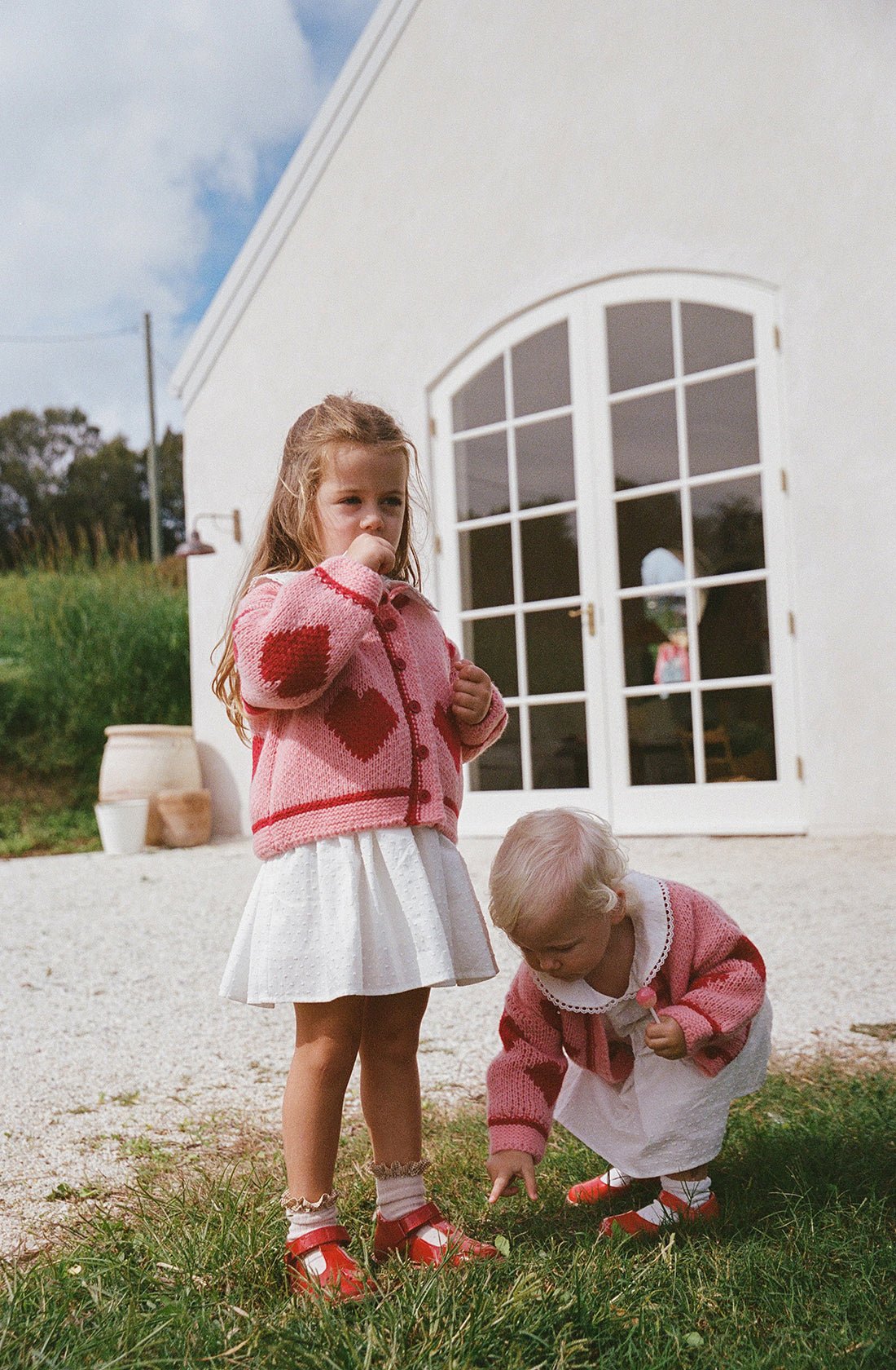 Two young children stand outside a building with large windows and a white door. They wear relaxed fit white skirts, Lover Knit Cardigan Mini from THE WHOLESOME STORE in pink and red heart patterns, and red shoes that are true to size. The older child stands while the younger one bends down, focused on something on the ground.