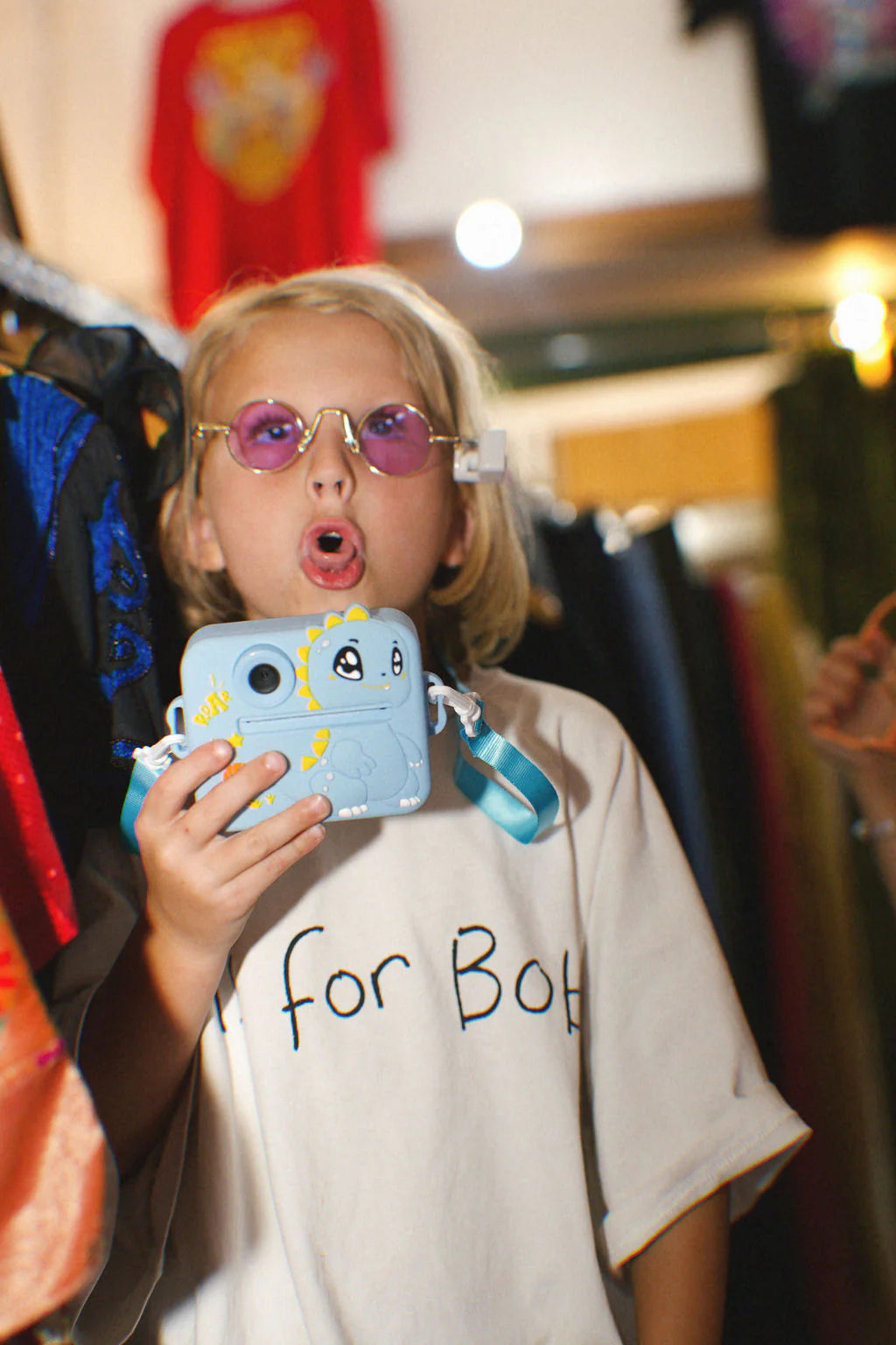 A blonde child wearing round pink-tinted glasses holds the Oh My Frankie ~ Oh My Snap Instant Print Camera Blue, featuring a cartoon character. They seem to be singing or speaking in a white shirt, with colorful clothes in the background—a perfect moment of creative play captured by OH MY FRANKIE.