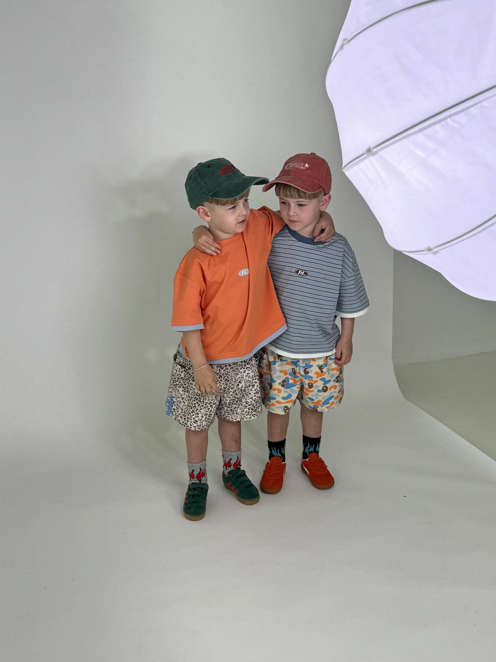 In a studio setting, two young boys stand close together, each wearing oversized outfits. They sport casual attire featuring hats, shorts, and sneakers. One boy has his arm comfortably draped over the other's shoulder, highlighting their friendship. Beside them is a large white light reflector. The boys are dressed in FRANCO'S DAD's PRE-ORDER Double Layer Tee Paw Paw.
