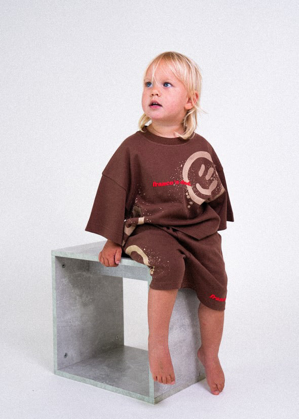 A blond child sits comfortably on a cube-shaped seat, wearing an oversized brown tee and FRANCO'S DAD Acid Dye Shorts Brown - Size 1, featuring a smiley face design. The stretch cotton set creates a cozy look against the plain white background.