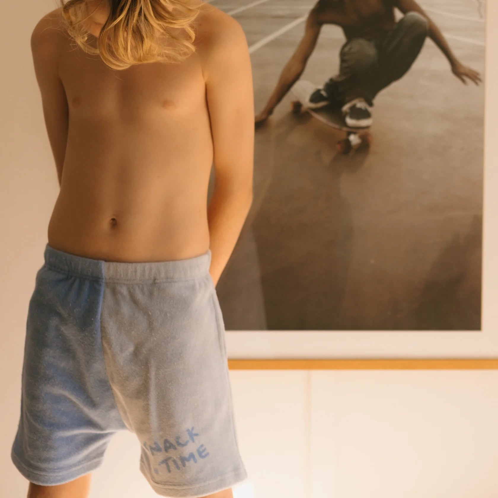 A person with long hair stands shirtless, wearing light blue and white Play Lounge Shorts Pool by GOLDEN CHILDREN that say "SNACK TIME." In the background, there is a large poster of someone skateboarding. The indoor setting is softly lit.