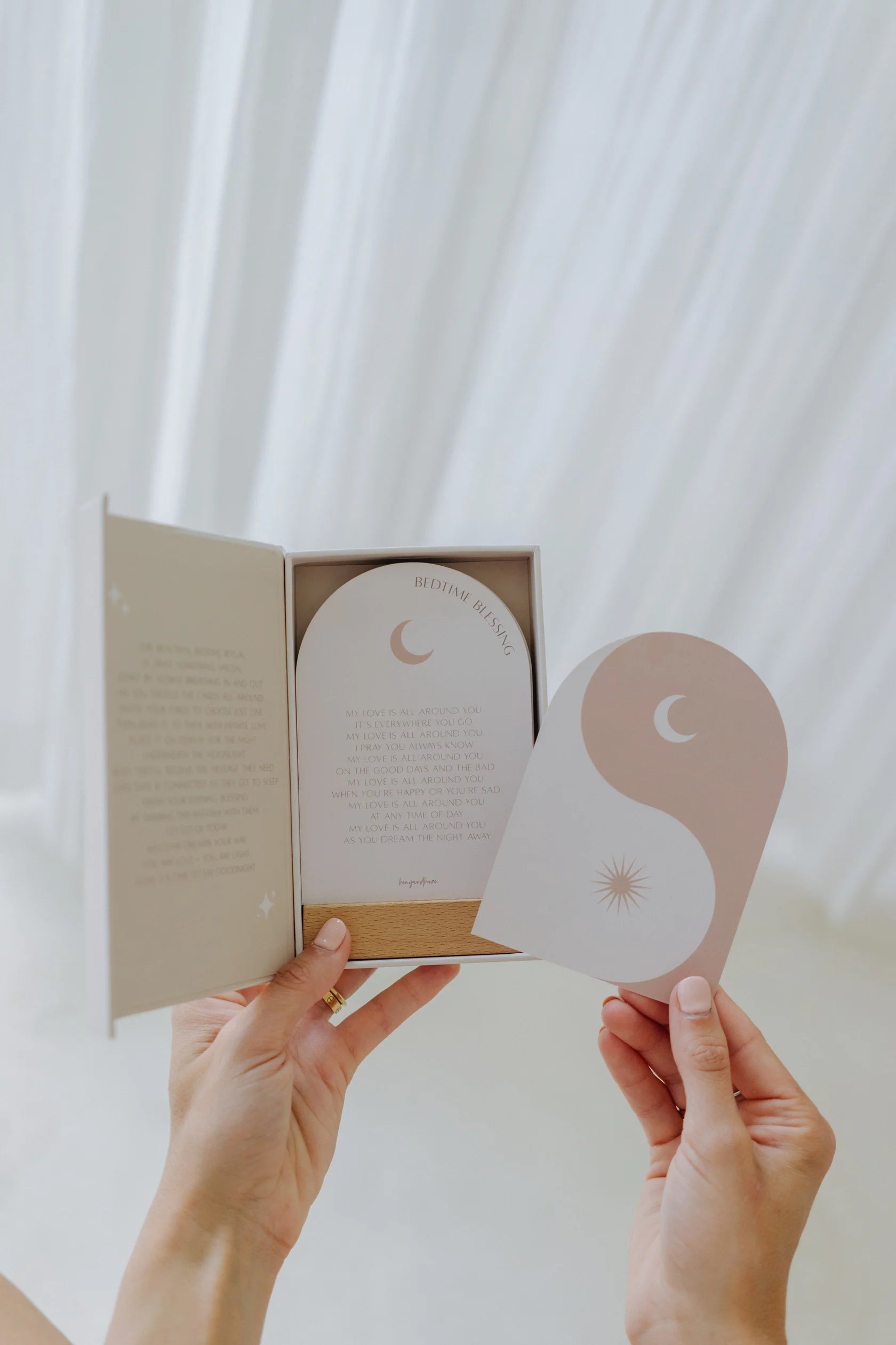 A person holds an opened box containing the Bedtime Blessings Deck by HENY AND HAZE, a set of bedtime reading cards adorned with soft pastel designs. One card features a crescent moon and star on a beige background, while another displays a yin-yang symbol. Ideal for kids' sleep challenges, these affirmation cards create serenity against the curtain backdrop.