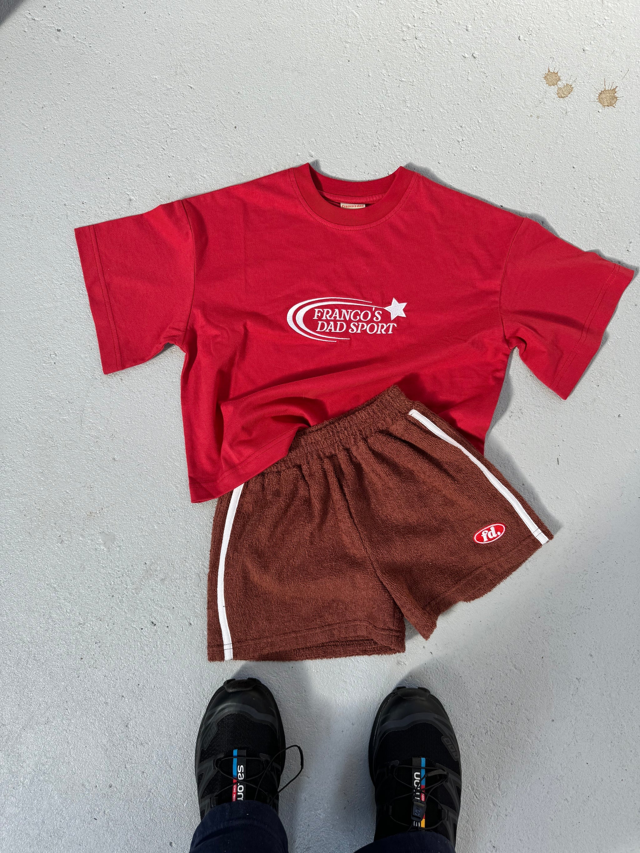 A red T-shirt featuring a white "Franco's Dad Sport" logo is styled with the PRE-ORDER Towel Shorts from FRANCO'S DAD. These brown athletic shorts feature white stripes and an emblem, making them an ideal summer staple. The look is completed with black sneakers, all standing out against a gray floor in this effortlessly stylish ensemble.