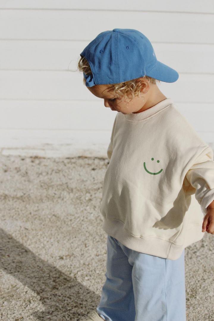 Young boy wearing the wave of life pullover in creme with a blue cap and pants looking down at the ground.