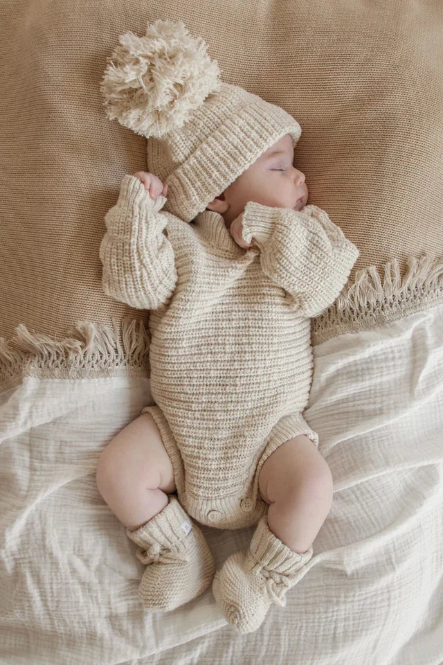 A sleeping baby is dressed in a cozy Heirloom Romper Honey from the ZIGGY LOU brand, paired with a knitted hat featuring a large pom-pom, lying on a soft, textured beige blanket. The romper, which includes a sweater, socks, and onesie in light cream from the Ziggy Lou Honey Signatures Collection, creates an overall warm and snug appearance.