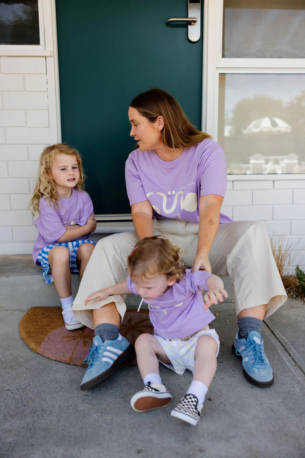 A woman in a Sunday Siblings Il Sole Tee in purple/cream sits on a doormat before a green door with two children, all in purple shirts. The cotton fabric adds comfort as she gazes at one child beside her, while the other relaxes on the ground ahead of her.