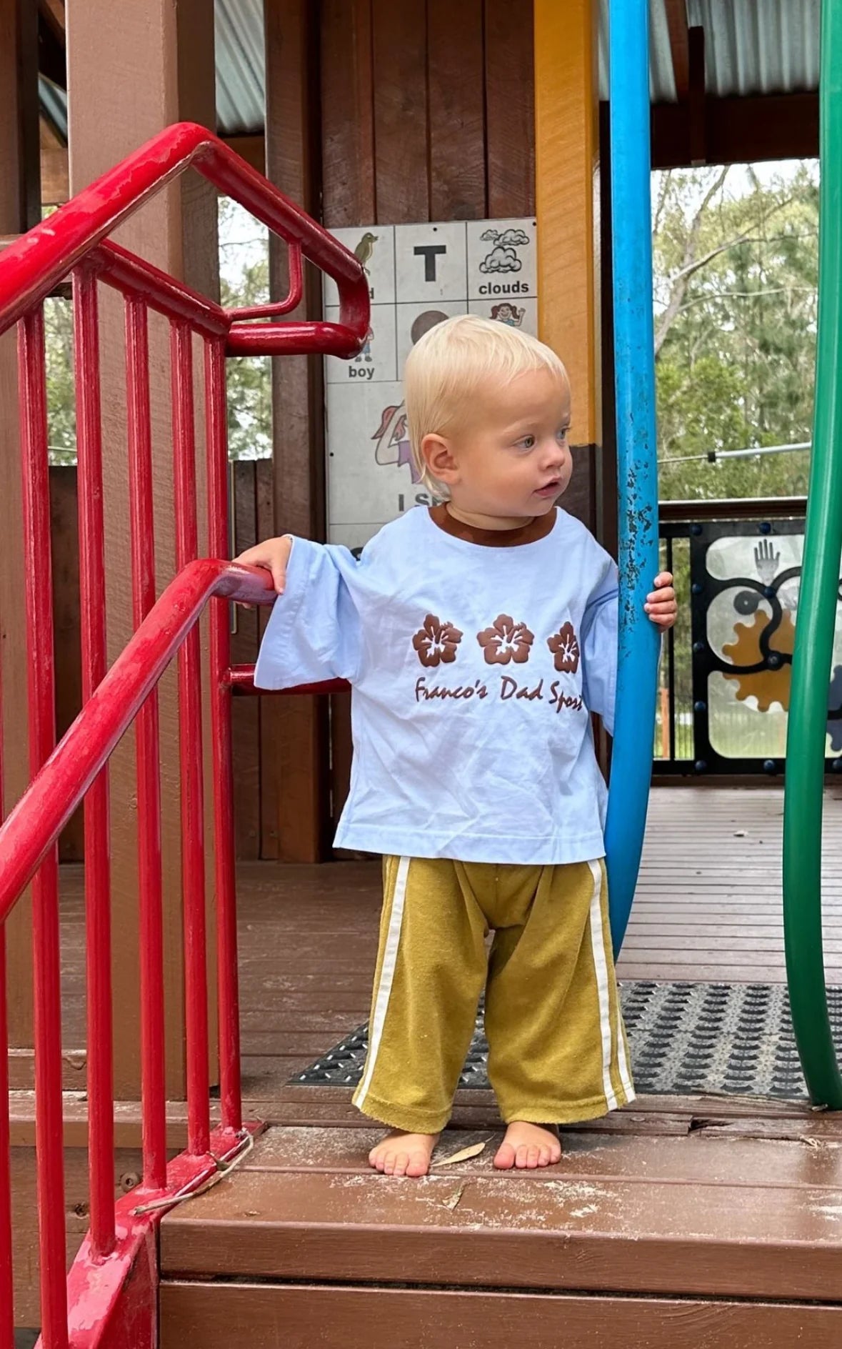 A young child with blonde hair stands barefoot on a playground structure, clutching a red railing and a blue pole. Dressed in an oversized PRE-ORDER Hibiscus Tee from FRANCO'S DAD, featuring a subtle design, and bright yellow pants, the child looks to the right. Trees and part of the play area are visible in the background.