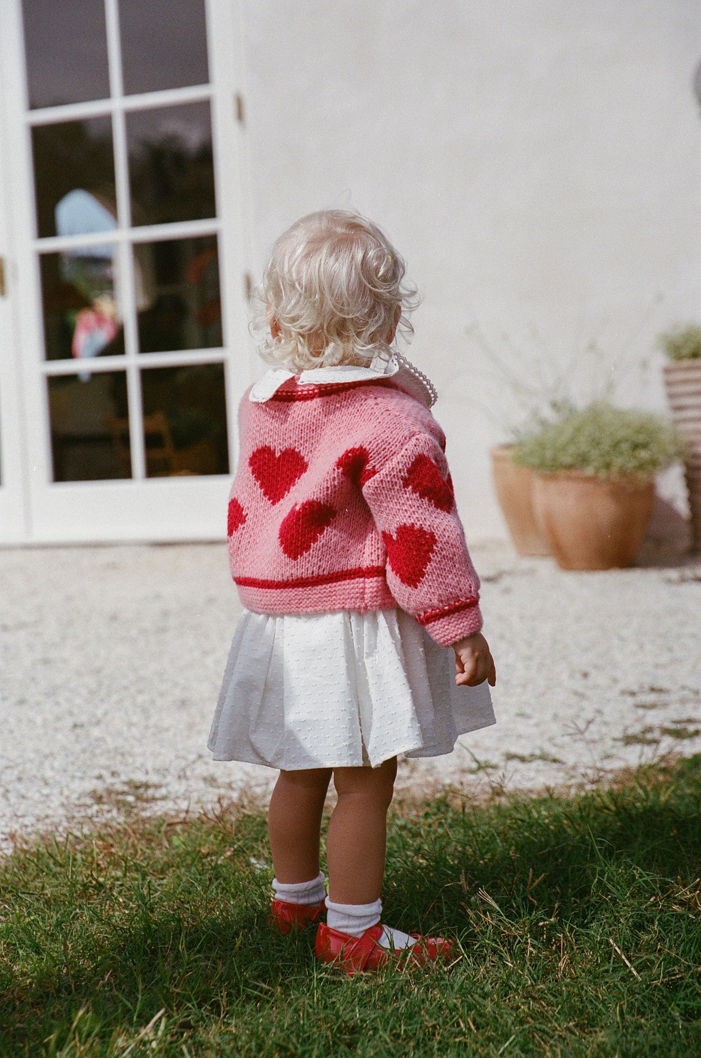 A young child with curly blonde hair wears THE WHOLESOME STORE’s Lover Knit Cardigan Mini—a relaxed-fit pink sweater with red hearts—over a white dress, paired with red shoes and white socks, standing on grass facing a glass door.