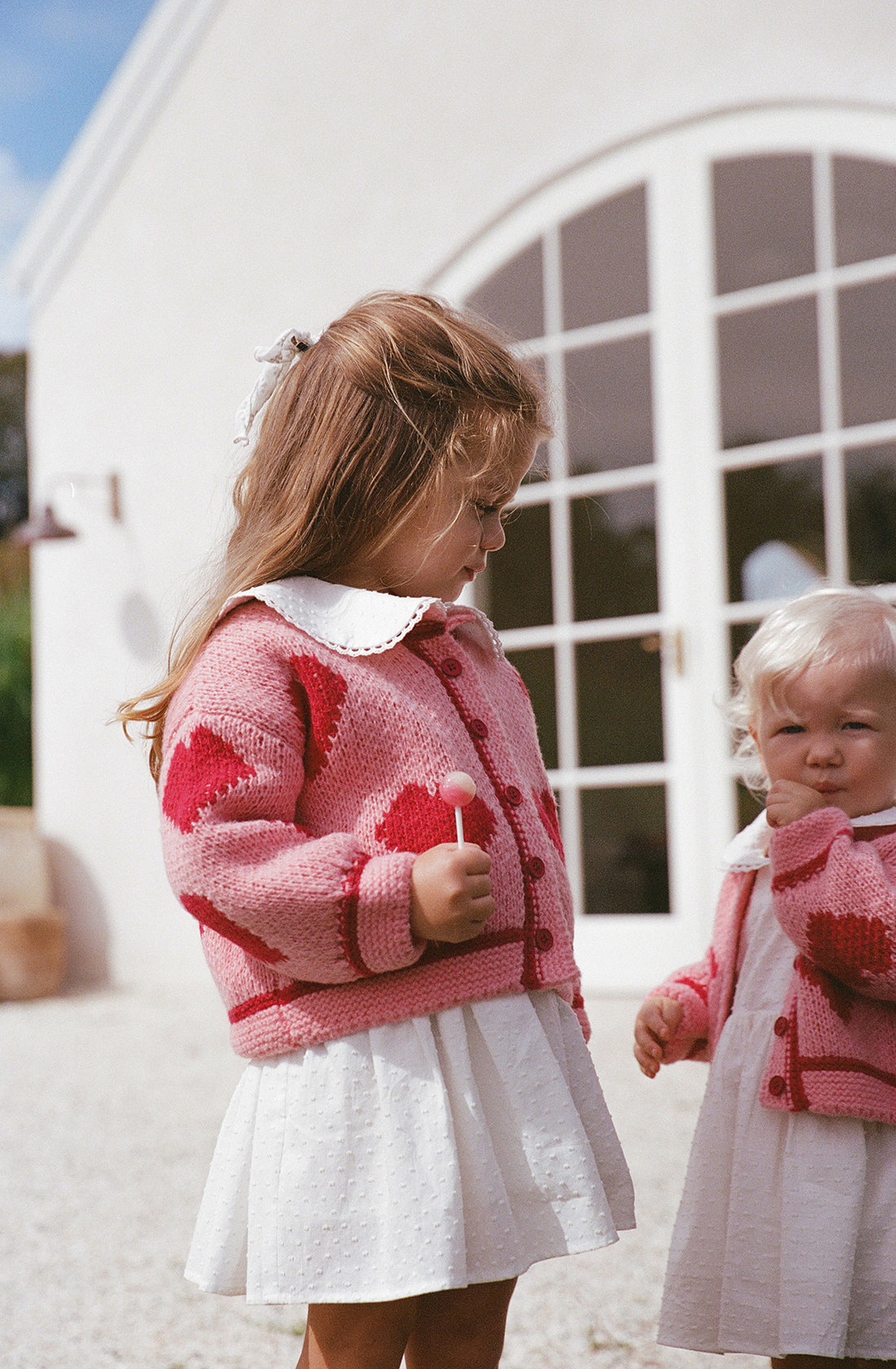 Two young girls stand in front of a white building with arched windows. The older girl wears THE WHOLESOME STORE's Lover Knit Cardigan Mini, which is pink with heart patterns, paired with a relaxed fit white dress, and holds a lollipop. The younger girl wears a similar outfit and looks at the camera while touching her chin. Both outfits are ethically made and true to size.