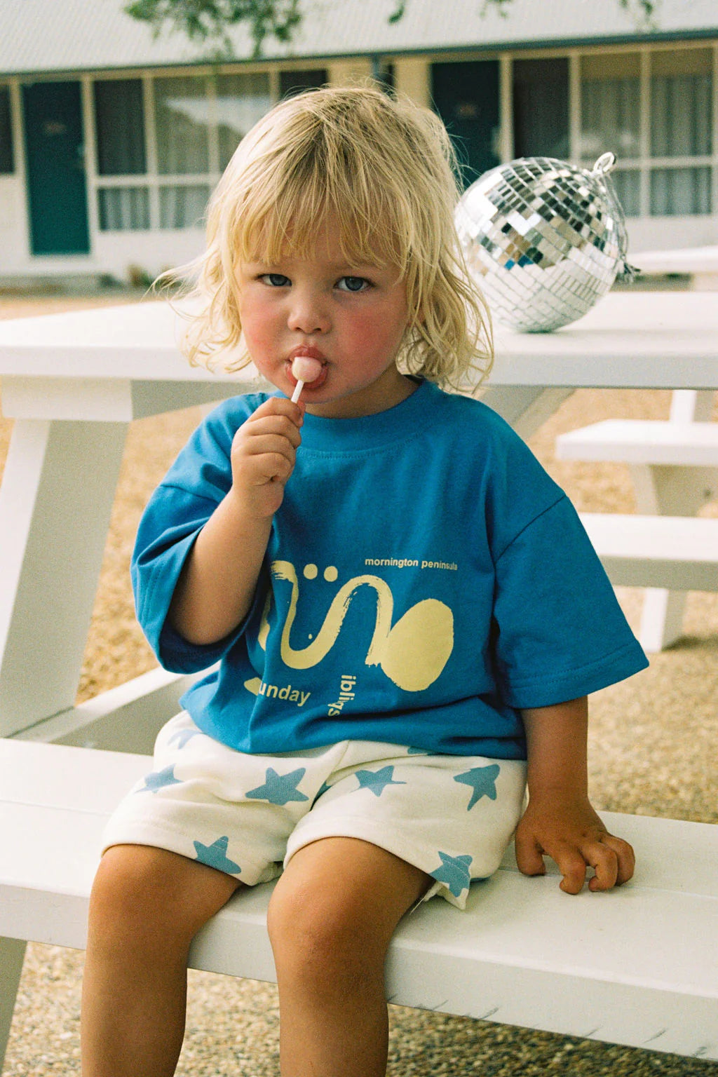 A young child with blonde hair sits on a white picnic table wearing an oversized Sunday Siblings ~ Il Sole Tee Blue/Yellow and star-patterned cotton shorts, licking a lollipop beside a disco ball, with a building having windows and a door in the background.