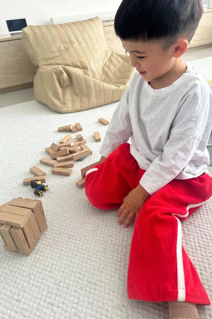 A young child in a white shirt and Darcy Racer Pants Red by TINY TROVE, featuring an elastic waistband, is sitting on a light-colored carpet. They are smiling at a disassembled Jenga game and a small toy figure nearby, with a beige cushion in the background adding to the cozy atmosphere.