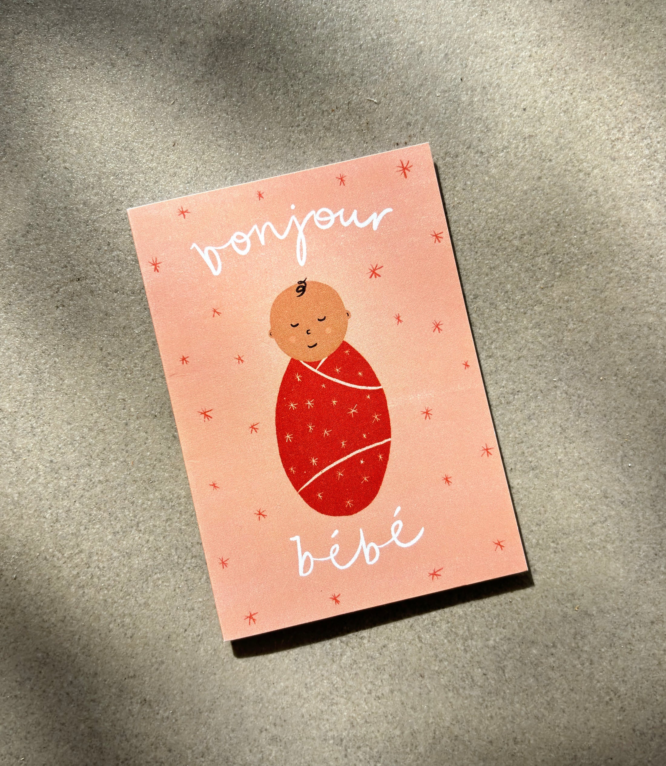 The Bonjour Bebe Pink Mini Card - New Baby from LAUREN SISSONS STUDIO features a charming illustration of a baby wrapped in a red swaddle on a textured, recycled paper surface. The card has a peach background adorned with small red stars and displays the text "bonjour bébé" in white cursive handwriting—an ideal choice for celebrating a baby shower.