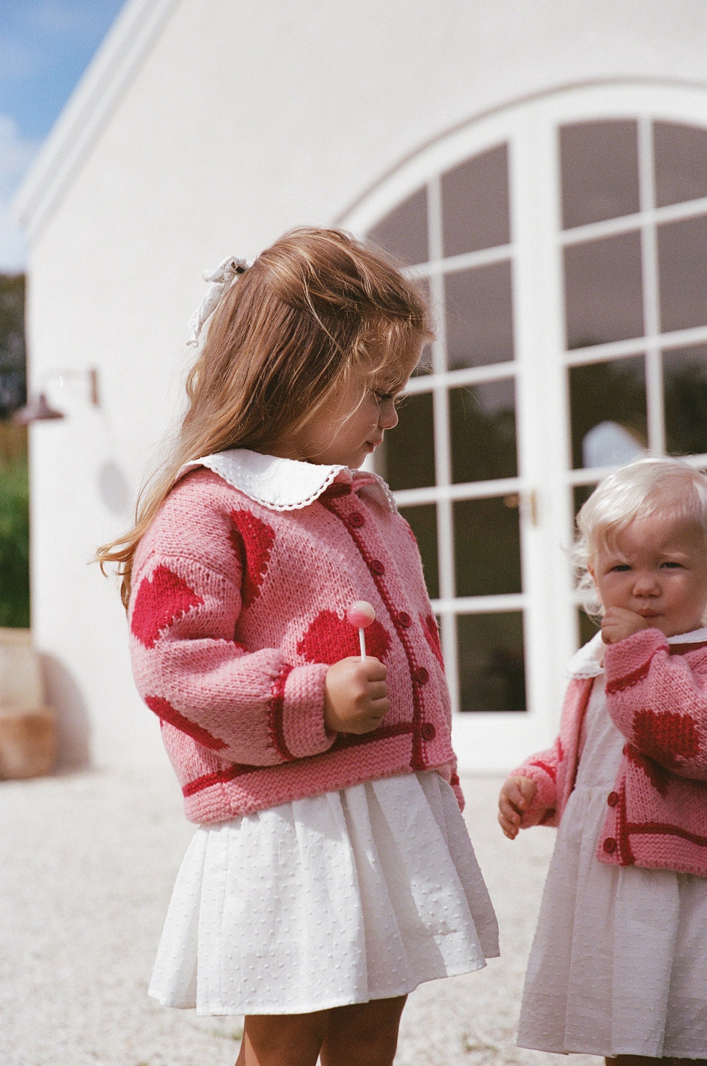Two young children stand outside a white building, wearing THE WHOLESOME STORE's Lover Knit Cardigan Mini in pink with red hearts over white dresses as they enjoy lollipops.