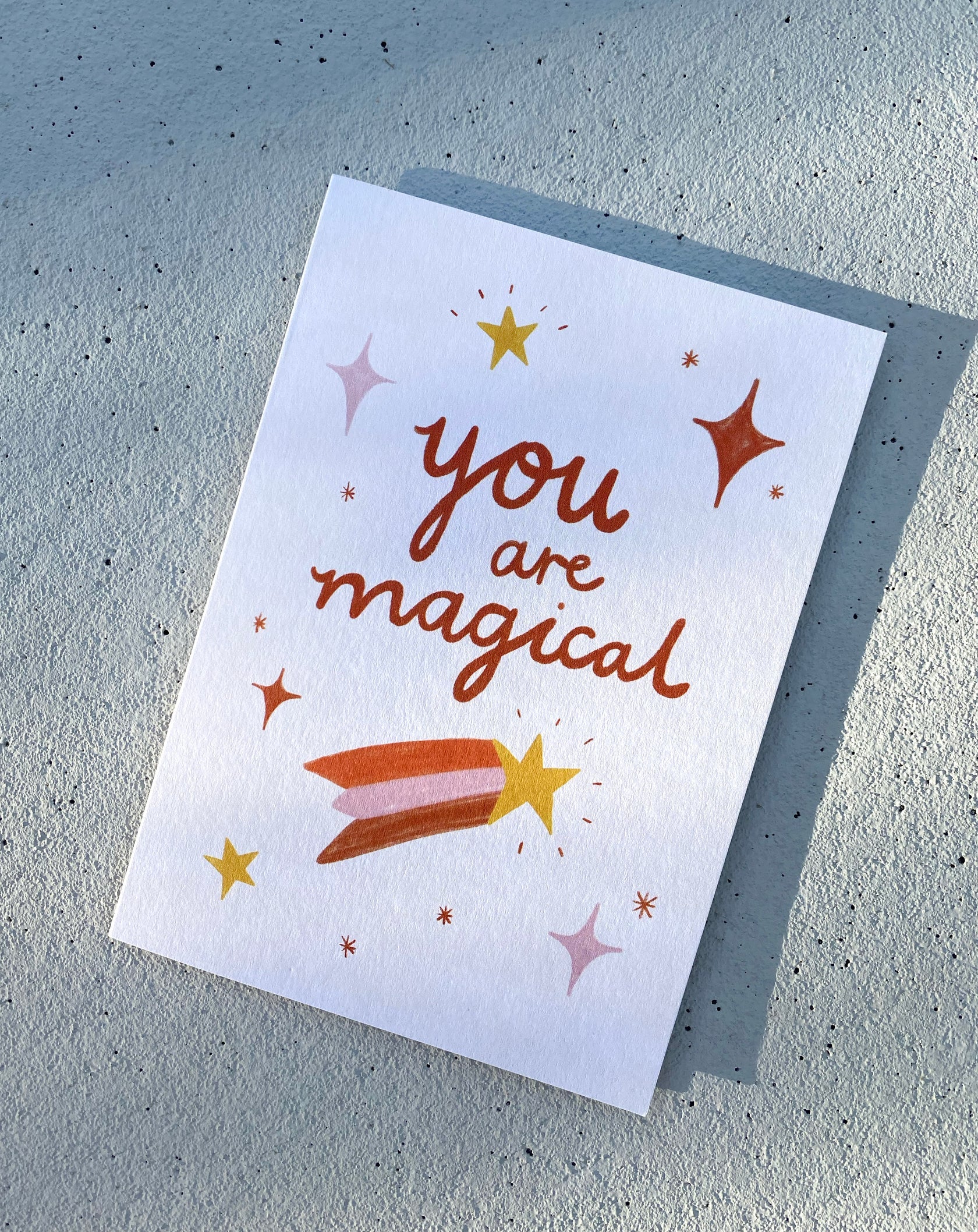 A LAUREN SISSONS STUDIO Greeting Card “You Are Magical” made from recycled paper rests on a light gray surface. It showcases the text "You are Magical" in red, adorned with several stars of different sizes and colors. Near the bottom of the card, a shooting star with a yellow head and red trail is depicted.