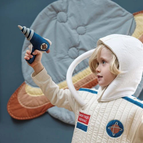 A child with blonde hair wears a white astronaut costume made from organic cotton, complete with a hood and a tube, while holding the FABELAB Dress Up Raygun Midnight Blue. The background features a wall hanging depicting a planet with rings, resembling Saturn—perfect for imaginative adventures.