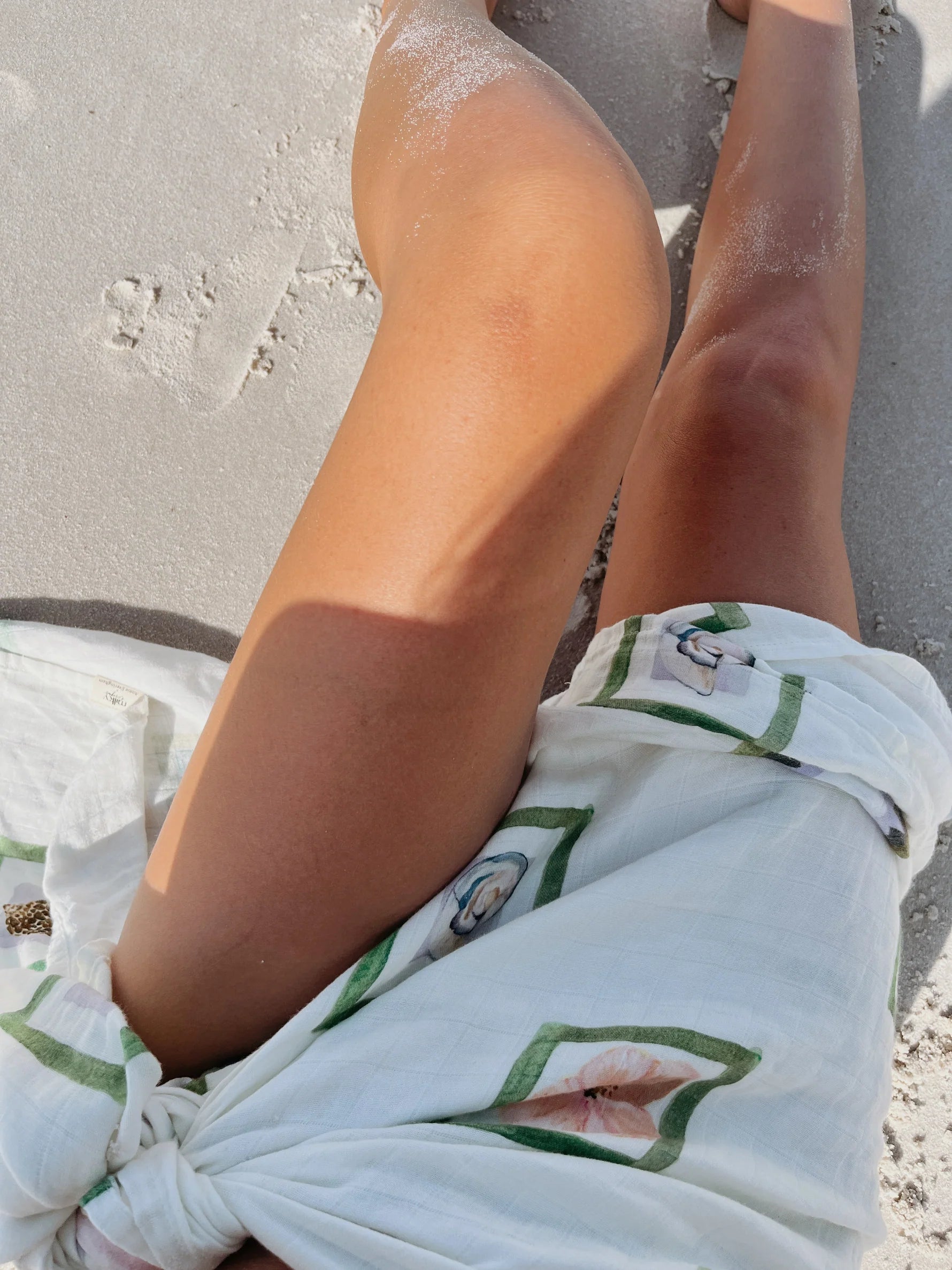 Lounging on a sandy beach, a person extends their legs while wrapped in the Milky Designs ~ Annie Wrap by MILKY DESIGNS, featuring Annie Everingham's green and floral patterns. Sand speckles the fabric and skin as sunlight adds a serene touch to the hand-painted design.