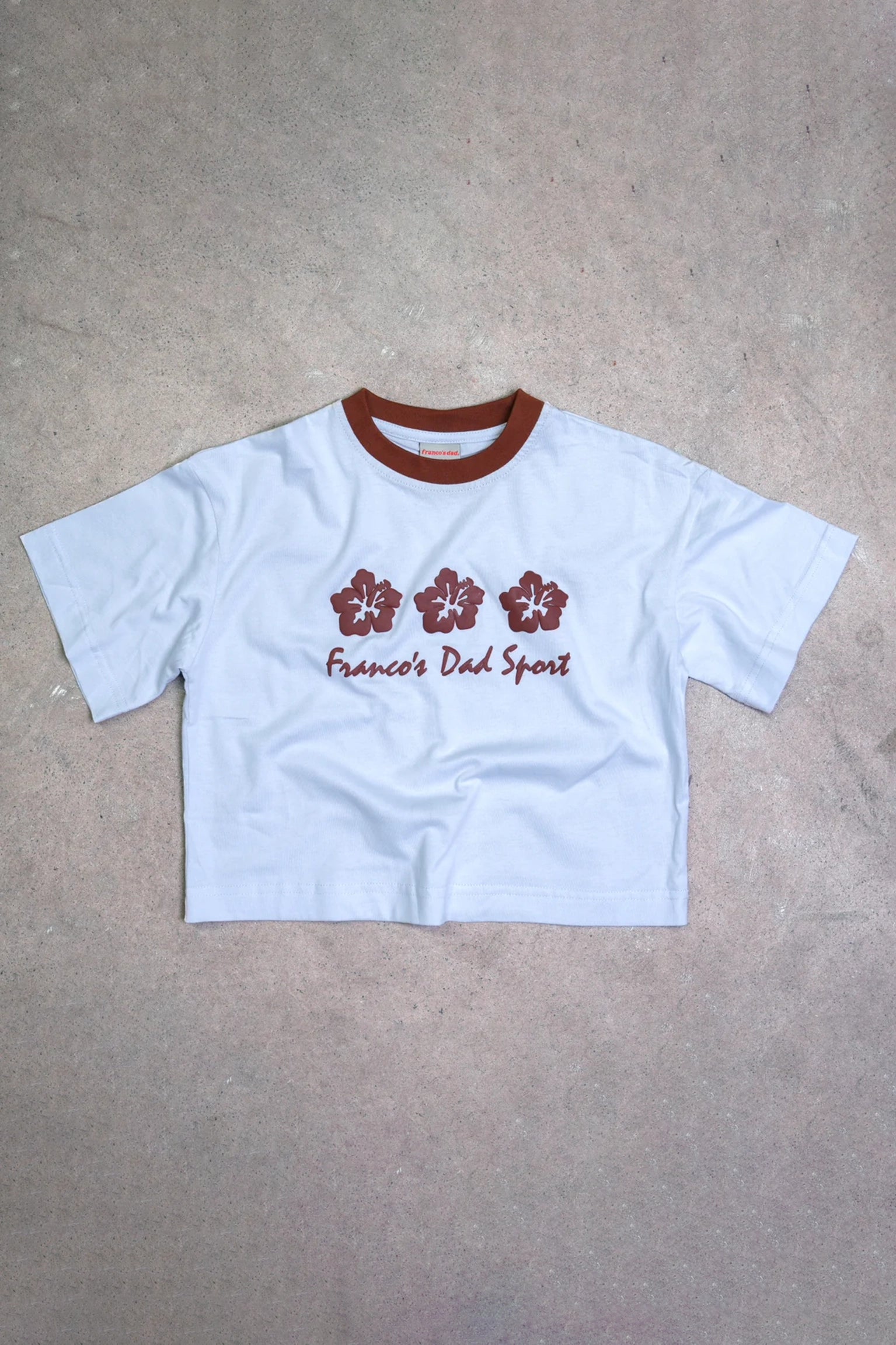 The PRE-ORDER Hibiscus Tee by FRANCO'S DAD is a cropped white T-shirt with a brown collar. It showcases three brown hibiscus flowers and features red text reading "Franco's Dad Sport" on the front, embodying a vintage 90's sports design vibe. The shirt is displayed flat against a beige background.