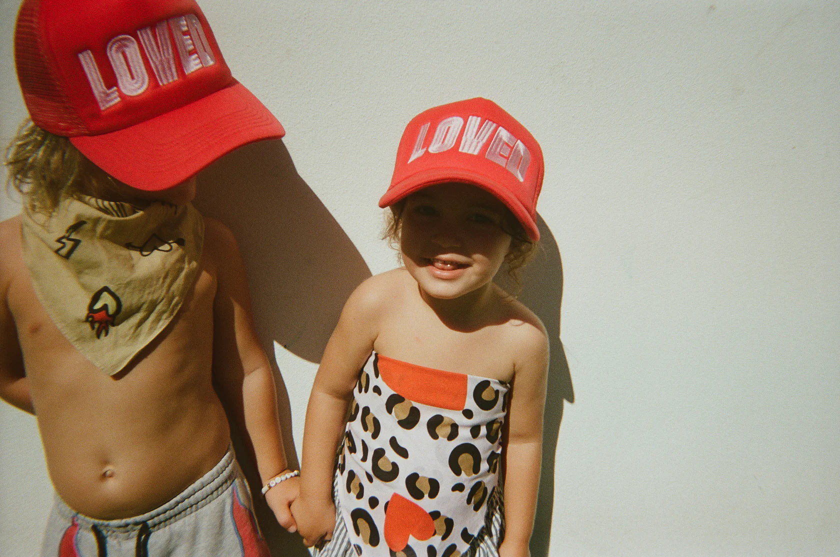 Two children standing against a white background, one older child and one younger child. Both are wearing "So Loved Trucker - Red" caps from TINY LOVE CLUB with the word "LOVER" on the front. The older child is shirtless with a beige bandana while the younger one is dressed in a leopard print outfit with red hearts and an adjustable back strap.