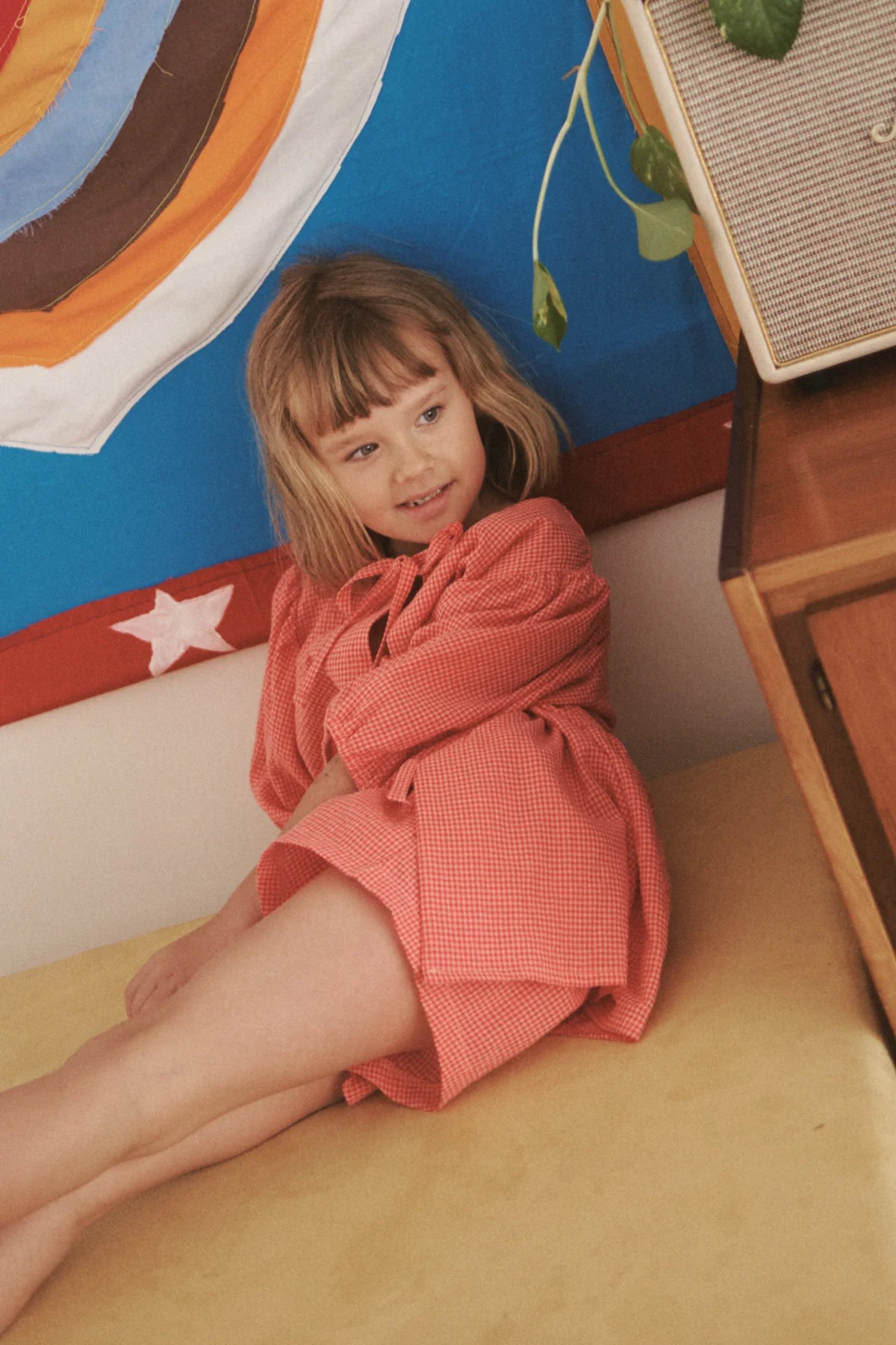 A young girl in LITTLE THE LABEL's Cami Shorts Cherry Gingham sits on a yellow surface, leaning against a colorful wall with a star design. She smiles, looking to the side, under hanging green leaves from a nearby plant.