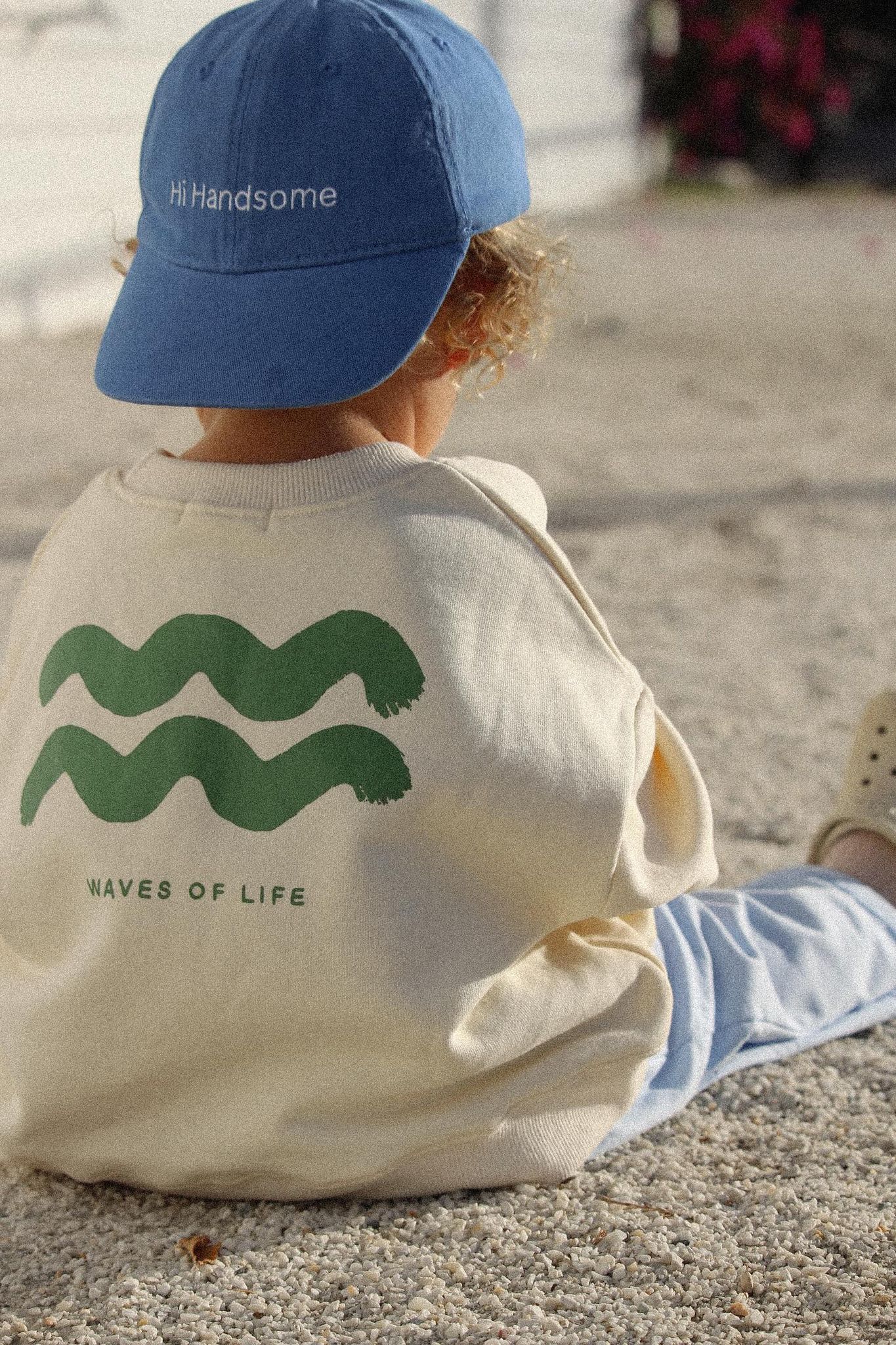 Young boy sitting down wearing the wave of life pullover in creme with a blue cap and pants.