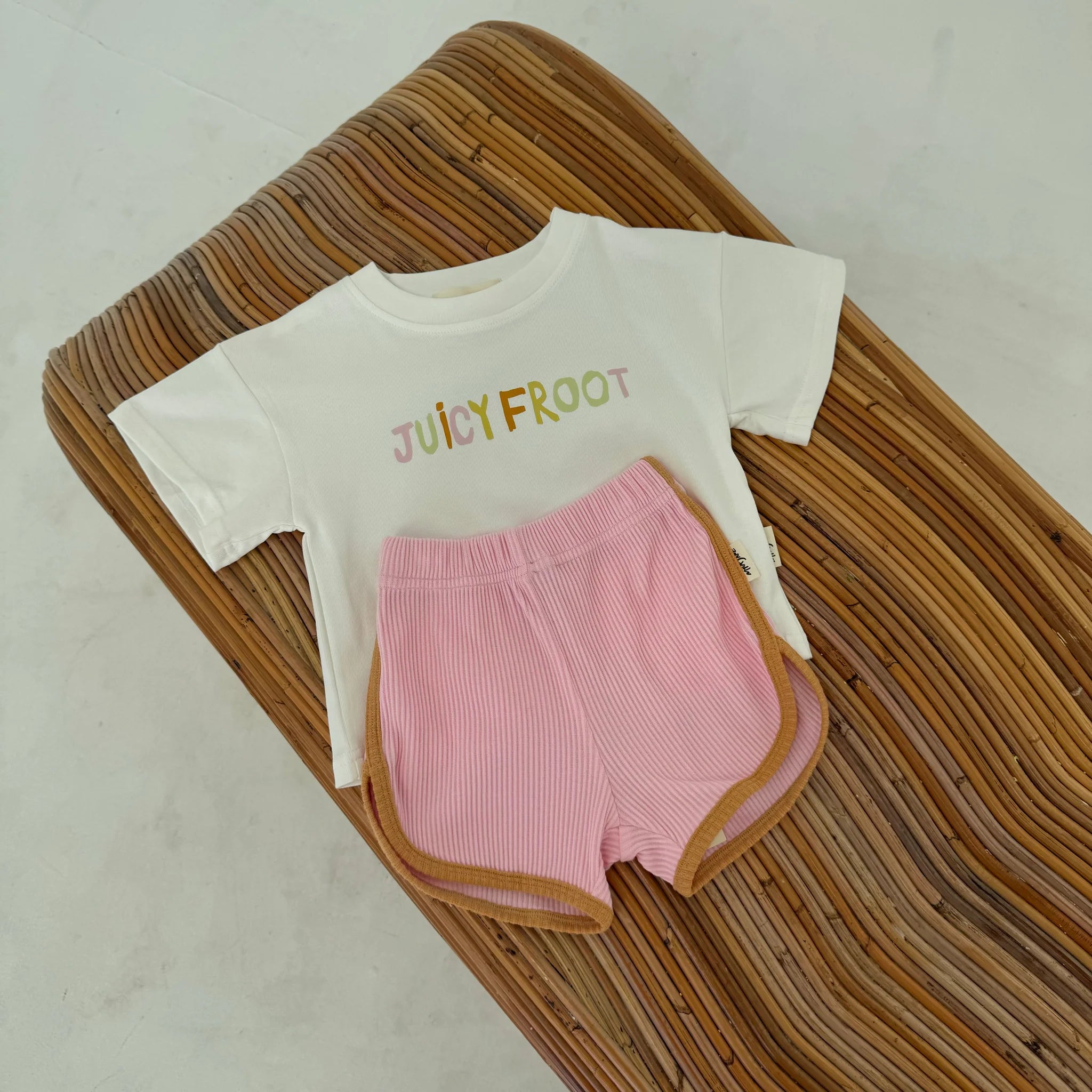 The product named "Contrast Rib Shorts Strawberry" by MYLA JANE, along with a white shirt, is displayed on a wicker surface. The ensemble features an oversized fit and is made from quality cotton rib for superior comfort.