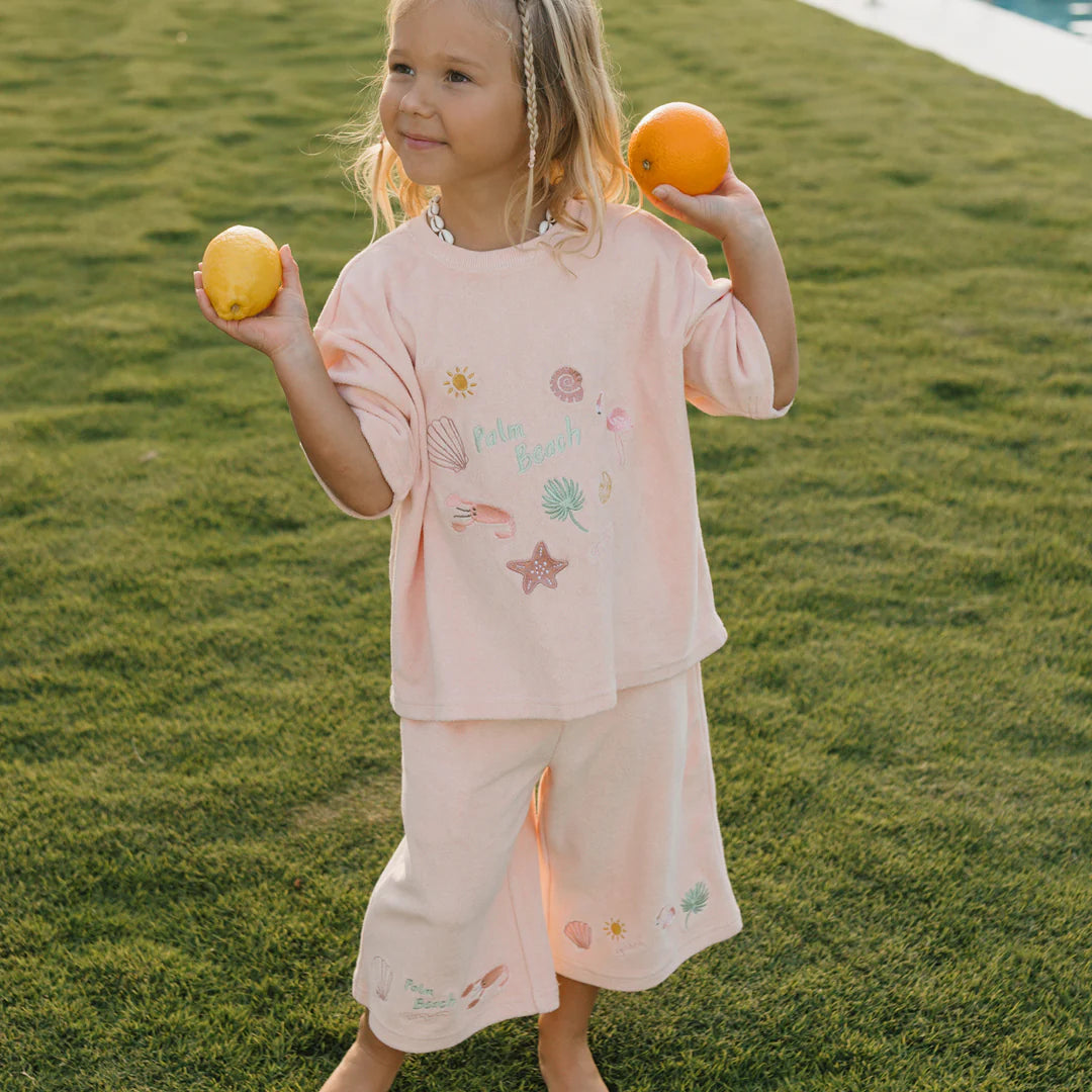 A young child stands on the grass, holding a lemon in one hand and an orange in the other. Dressed in a GOLDEN CHILDREN Palm Beach Mid Sleeve Tee Terry Towel Flamingo Pink, they pose happily near the pool in the background.