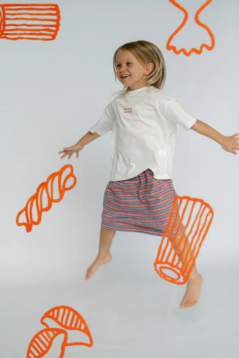 A barefoot child, with blonde hair, joyfully jumps in the air against a plain backdrop. The child is wearing an oversized fit white "Pasta Tee" from LENN LABEL and a striped knee-length skirt. Abstract red-orange line drawings of various whimsical shapes surround them.