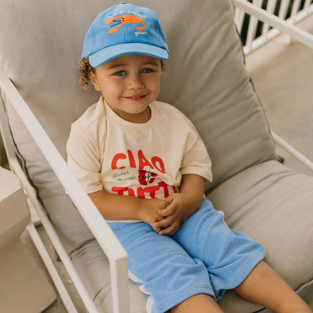 A young child sits comfortably on a cushioned chair, wearing a light-colored T-shirt with red lettering, light blue pants, and the GOLDEN CHILDREN Lobster Cap Deep Sea Blue featuring an orange lobster design and an adjustable brass clasp. The child smiles warmly at the camera.