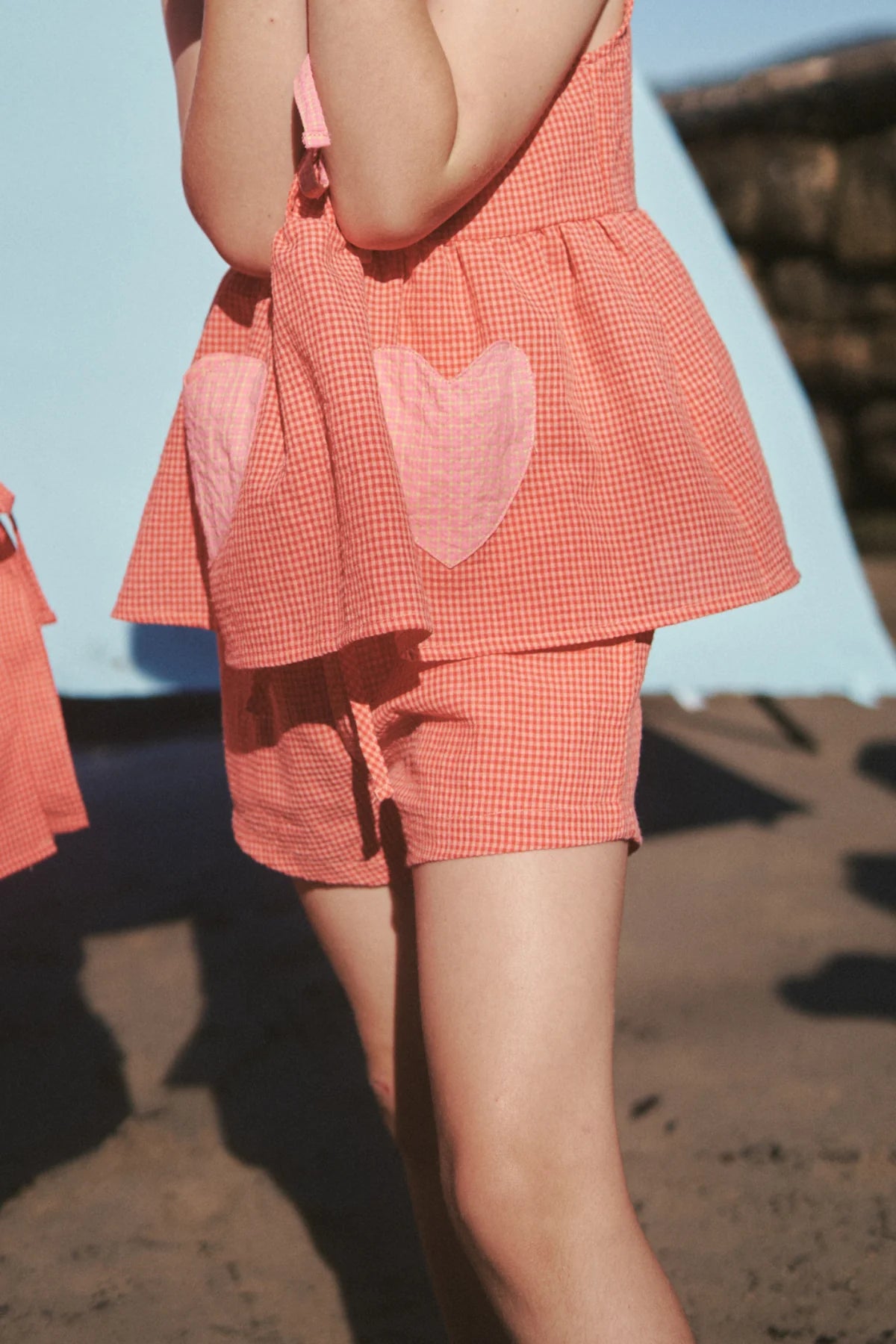 A person in LITTLE THE LABEL's Cami Shorts Cherry Gingham, featuring pink heart-shaped pockets, stands on a sandy surface. The seersucker cotton outfit includes a sleeveless top and shorts, set against a light blue backdrop.