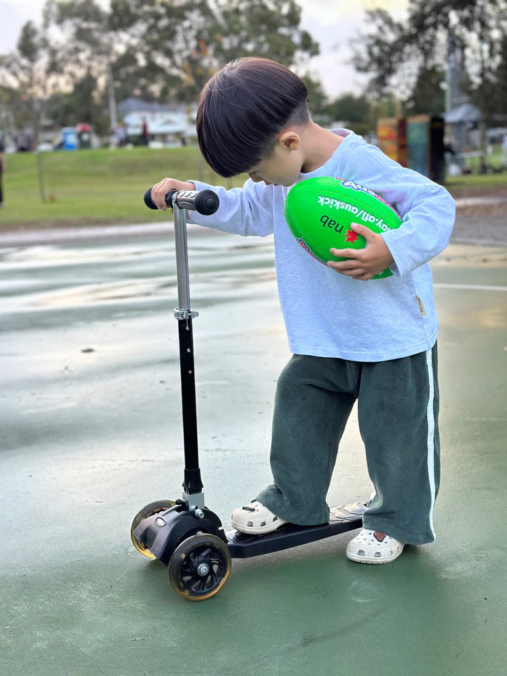 A young child wearing a light blue long-sleeve shirt and TINY TROVE's Darcy Racer Pants Moss stands on a scooter in a park. The child is holding a green ball and looking down at the scooter. Trees and other greenery are visible in the background.