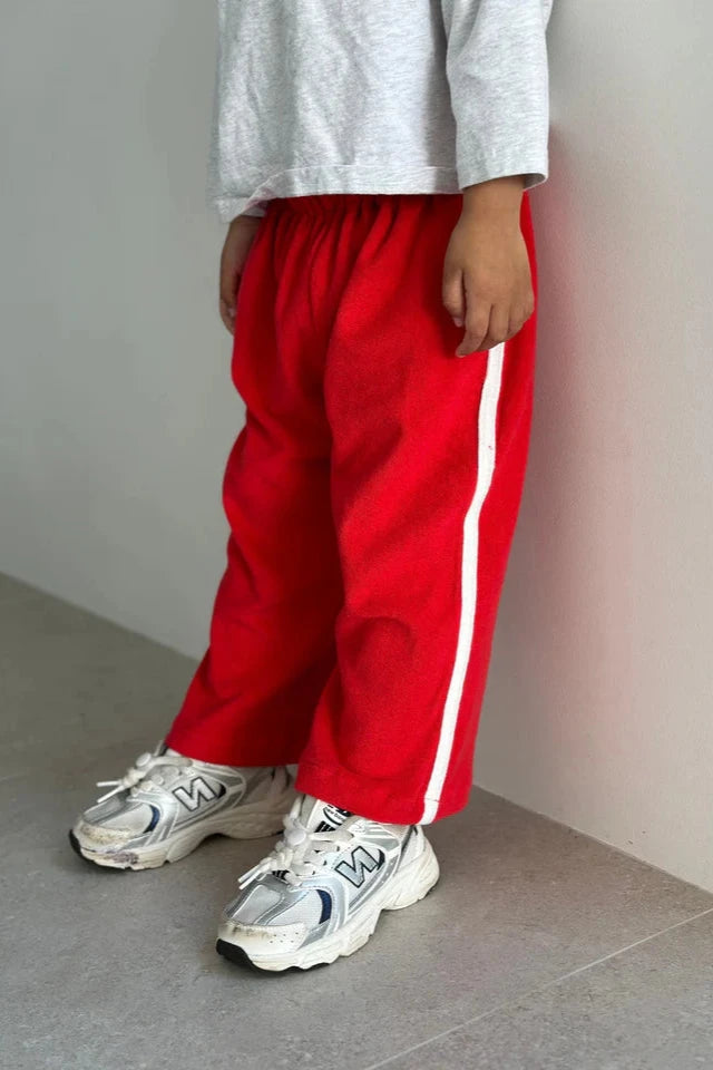 A child stands against a wall wearing TINY TROVE's Darcy Racer Pants in bright red, featuring an elastic waistband and a white stripe down the side, paired with a light gray long-sleeve shirt and white sneakers. The light-colored floor enhances the casual and comfortable vibe of the outfit.