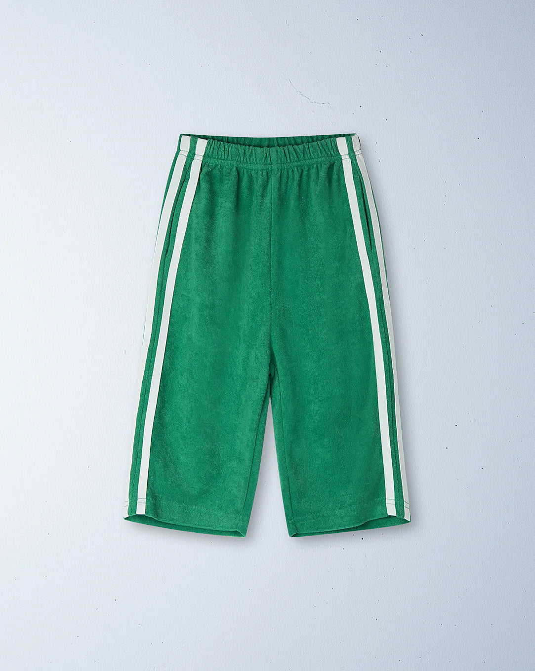 The Ziggy Zaza ~ Twin Stripe Terry Pant Verde by ZIGGY ZAZA are green knee-length shorts featuring an elastic waistband and dual vertical white stripes on each side, set against a plain light blue background. Made from 100% terry cotton.