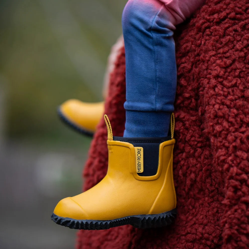 A child in blue pants and Merry People ~ Bobbi Kids Gumboot Mustard Yellow sits on a red textured blanket. The bright yellow boots, with vegan arch support and black soles featuring a small label, are 100% waterproof. The background is blurred and green.