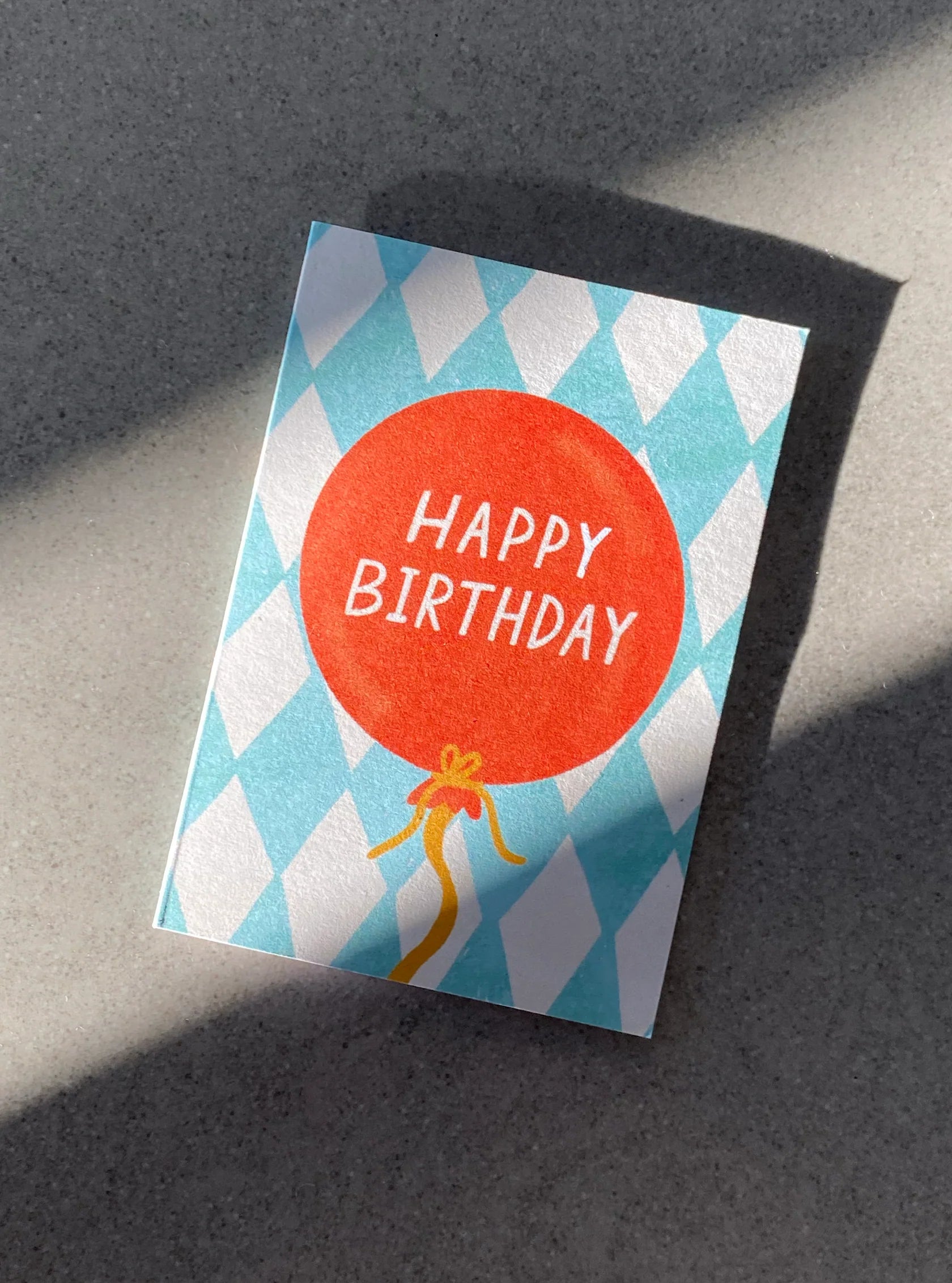 The Birthday Balloon Mini Card by LAUREN SISSONS STUDIO showcases an orange balloon and the words "Happy Birthday" in white against a blue and white diamond-patterned background. Made from recycled paper, this eco-friendly card is displayed on a gray surface with shadows.