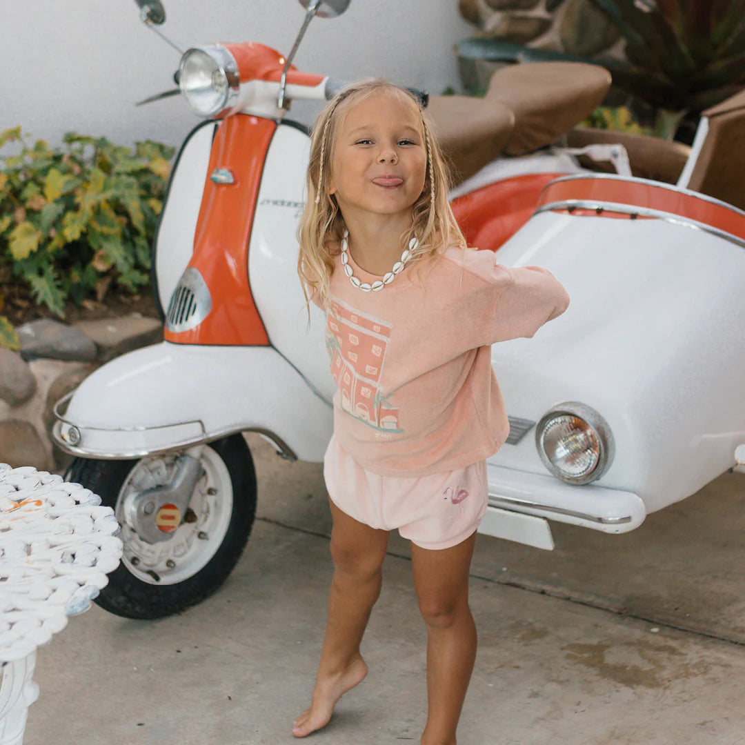 A young girl beams with charm as she poses in front of a vintage red and white scooter with a sidecar. She's wearing her GOLDEN CHILDREN Room Service Mid Sleeve Tee Terry Towel Spanish Villa, along with matching shorts and a shell necklace. The lush outdoor setting mirrors the vibrant atmosphere of the Colony Hotel.