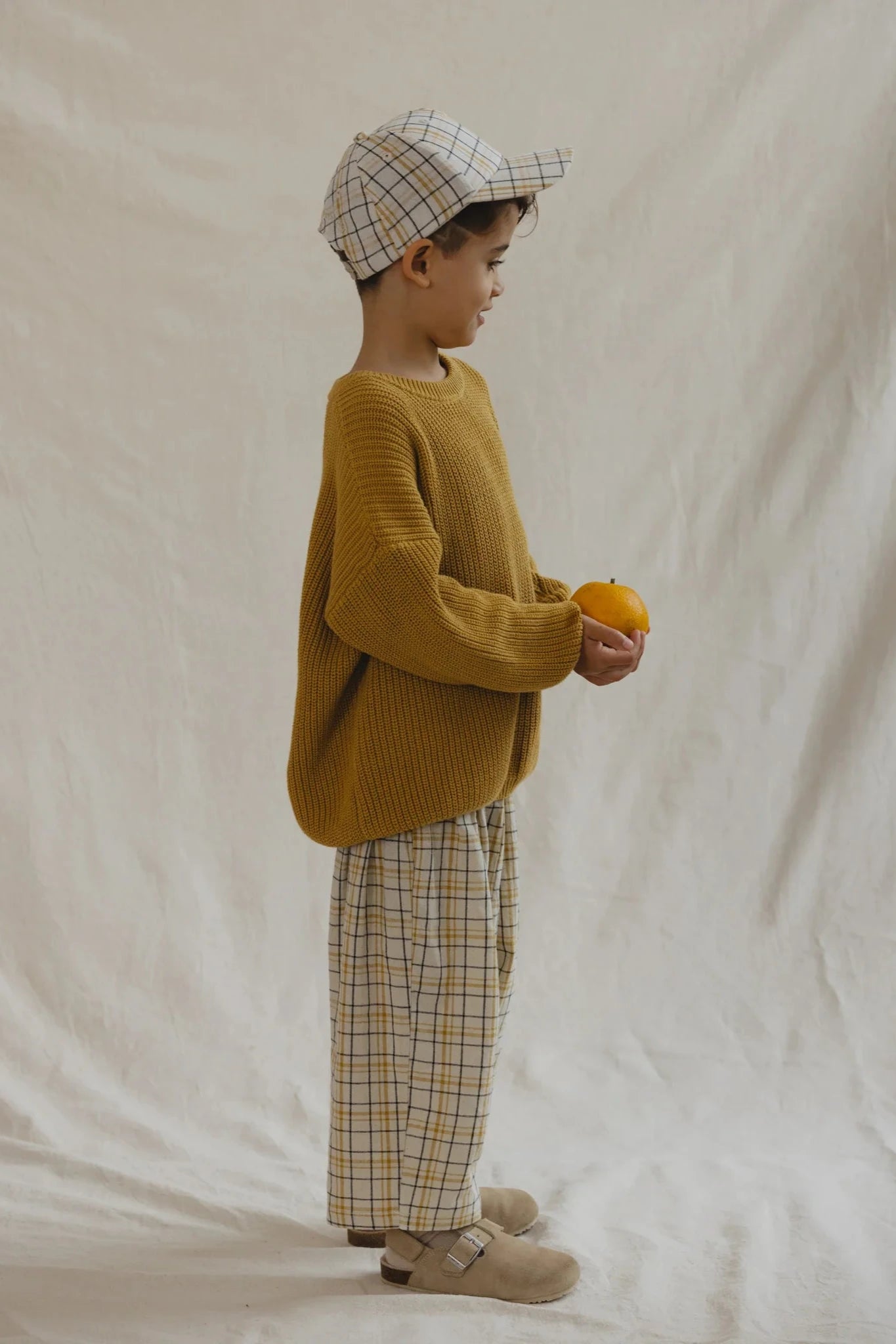 A young child wearing a yellow knit sweater, durable and practical 100% cotton Rio Pant Picnic by VALENCIA BYRON BAY, a matching plaid cap, and beige shoes stands in profile against a light, textured background while holding an orange in both hands.