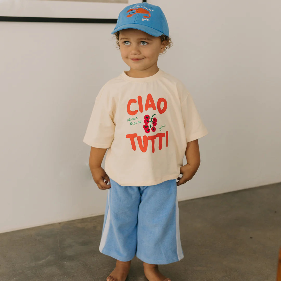 A young child stands indoors, dressed in a light-colored "Ciao Tutti" t-shirt and blue pants. They are wearing the stylish Lobster Cap in Deep Sea Blue from GOLDEN CHILDREN, featuring an adjustable brass clasp. The child has curly hair and is smiling against a plain, light-colored wall with flooring partially visible.