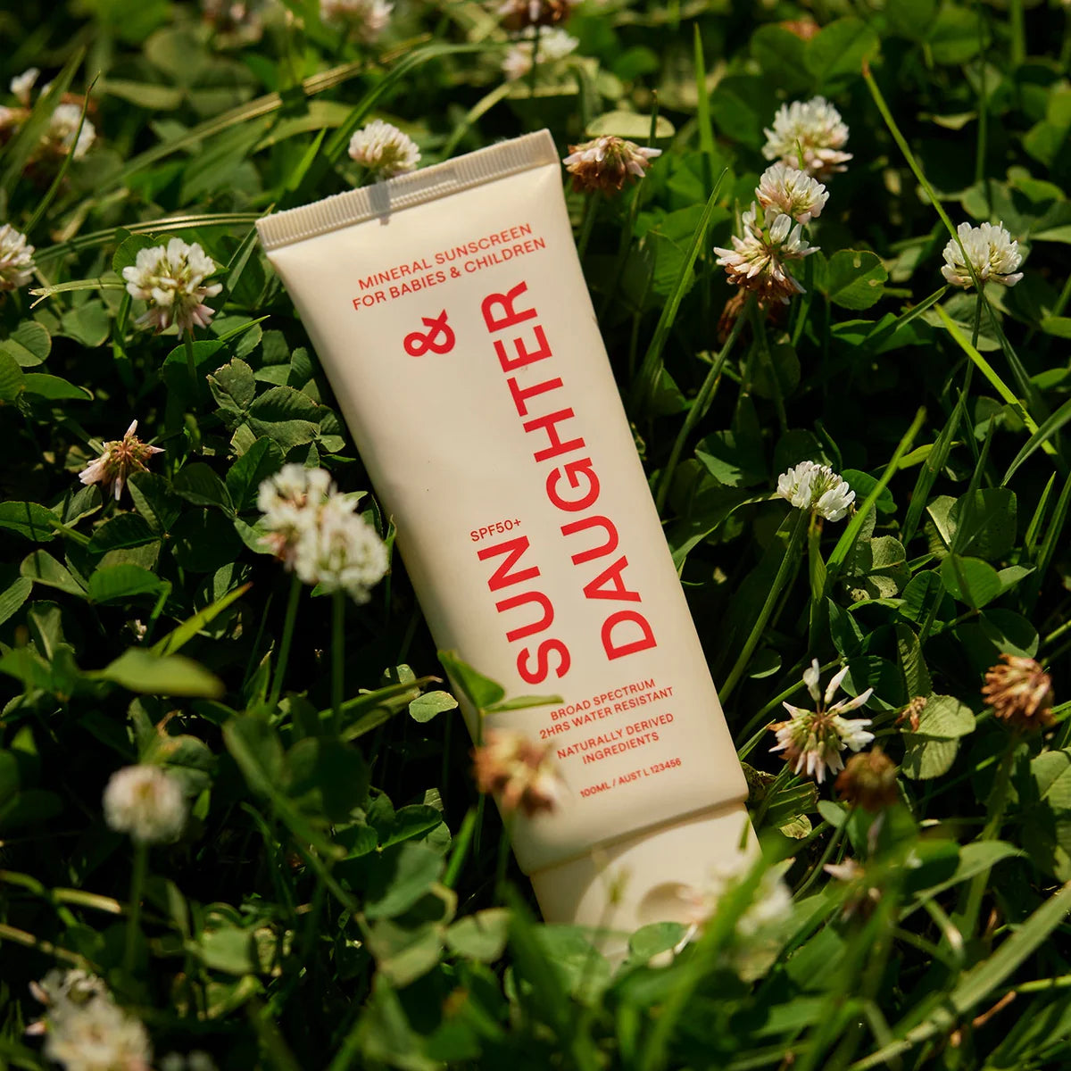 A tube of SUN & DAUGHTER SPF50+ mineral sunscreen, known for being reef-friendly and designed for babies and children, rests on the grass among clover flowers, providing broad-spectrum UVA/UVB protection.