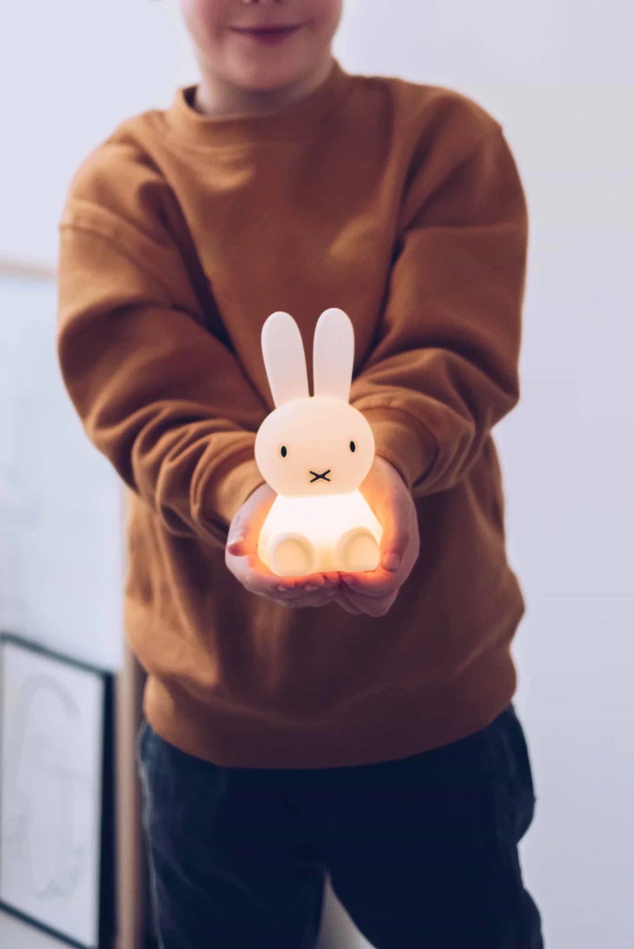 A child, visible from the shoulders down, is holding a lit MR MARIA Bundle of Light - Miffy night light made of soft silicone with both hands. The child, dressed in a brown sweater, extends their arms forward to offer the glowing toy. The background is softly blurred.