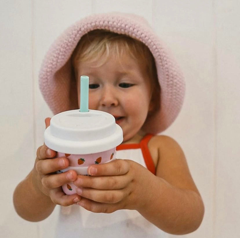 A small toddler holding a reusable babycino 120ml cup with a blue straw and white lid. The cup has strawberries scattered over the cup on a pink background.