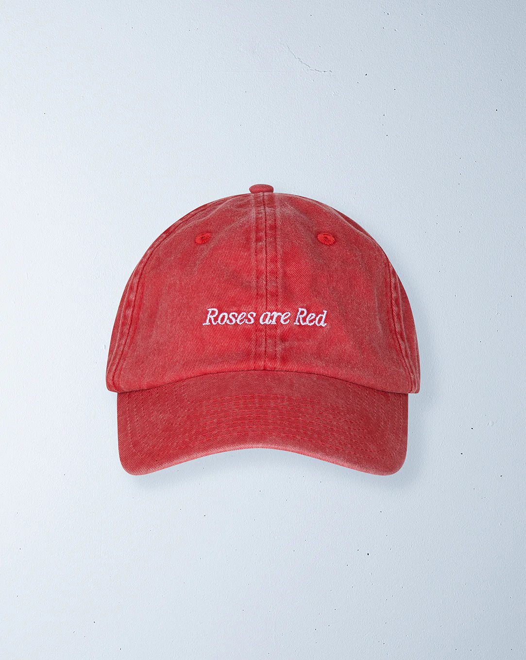 The Ziggy Zaza ~ Roses Are Red Cap by ZIGGY ZAZA is a washed red cotton canvas baseball cap with white "Roses are Red" embroidery. It features an adjustable brass clasp, six panels, a button on top, and a vintage texture against a light gray background.