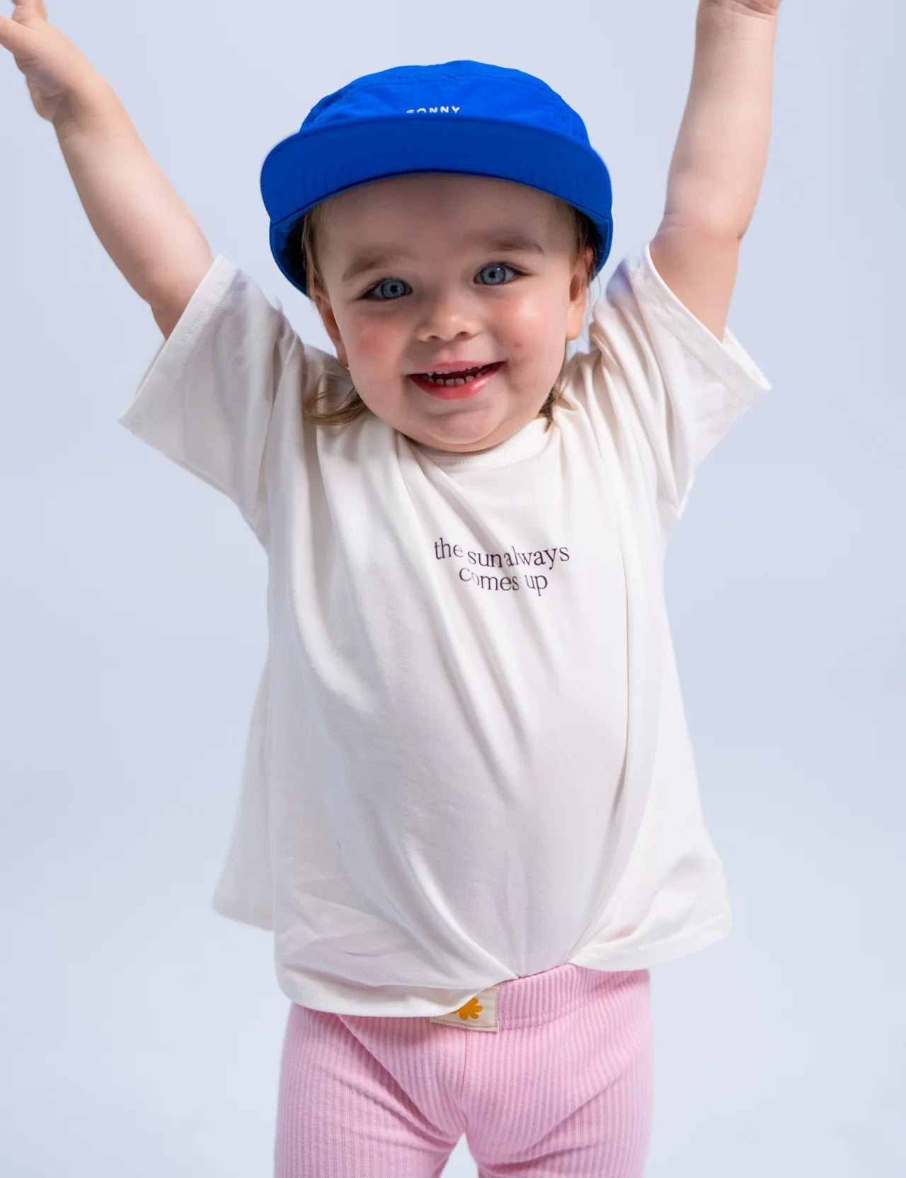 A joyful child wearing the Summer Club Cap in Electric by SONNY LABEL, a white T-shirt that reads "the sun always comes up," and pink pants raises their arms in excitement against a plain background.