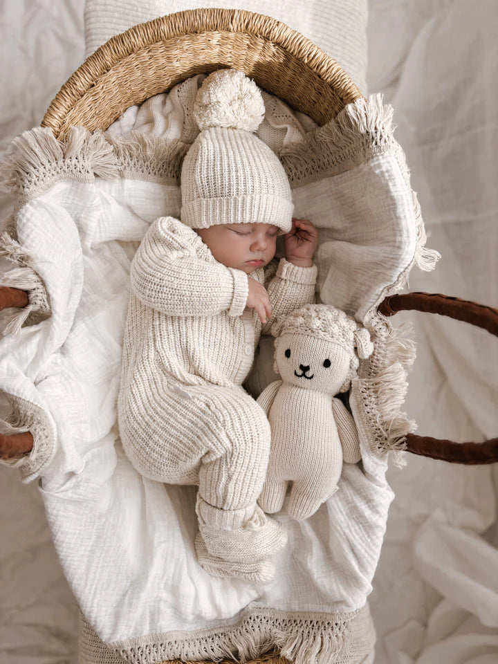 A sleeping baby dressed in ZIGGY LOU's Classic Knit Romper Honey, a cream-colored winter outfit with a matching hat, lies in a woven bassinet. The baby is cuddling a knit teddy bear toy that also wears a matching cream-colored outfit. The bassinet is lined with soft, textured cotton cloth.