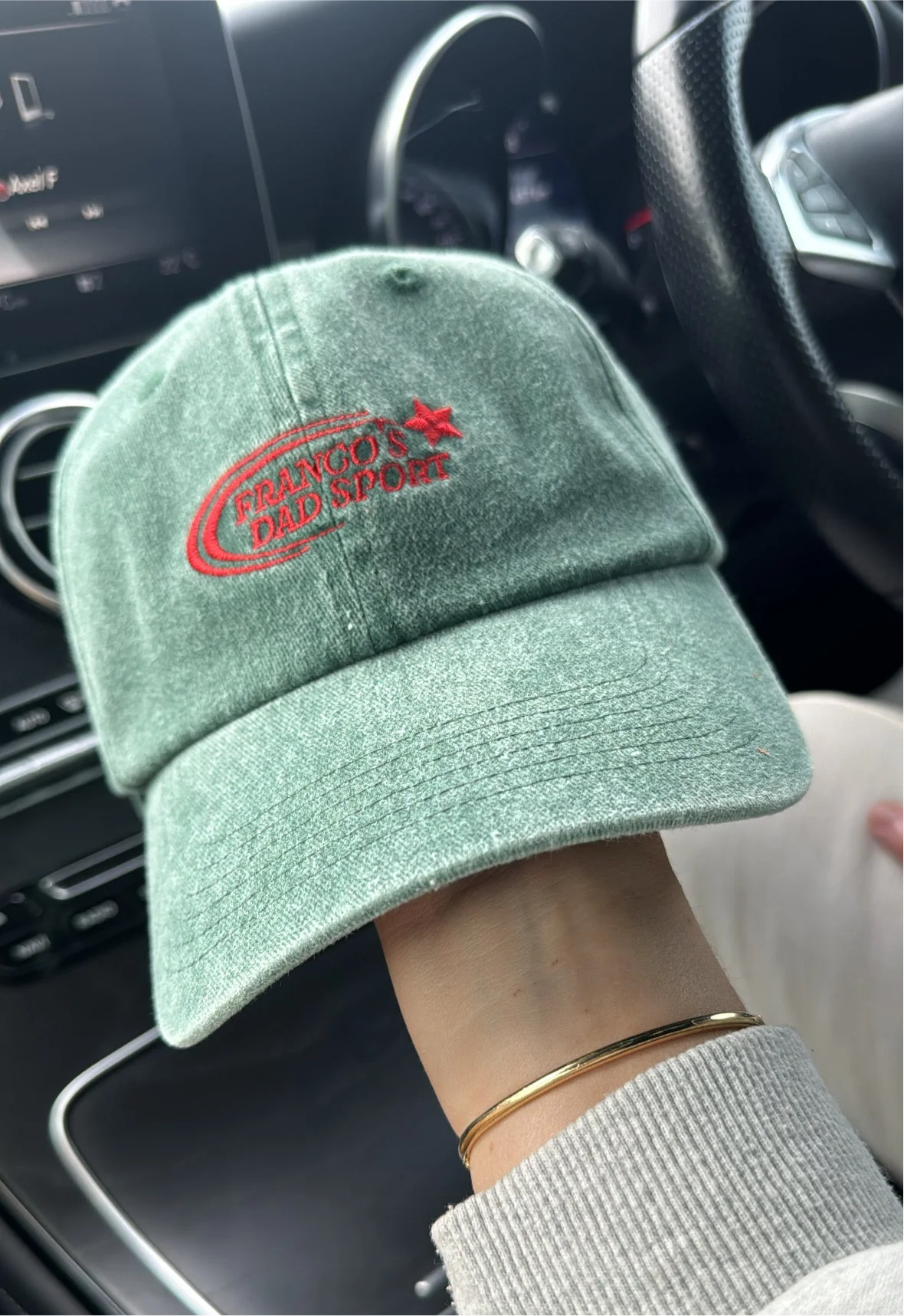 A hand holds a PRE-ORDER Sports Cap Green from FRANCO'S DAD, featuring red embroidery that reads "FRANCO'S DAD SPQR." The cap is displayed inside a car, with glimpses of the steering wheel and dashboard in the background. The person holding the cap is also wearing a bracelet.