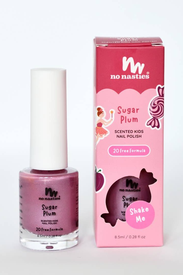 A bottle of "Sugar Plum" Water Based Scented Scratch Off Kids Nail Polish by NO NASTIES is displayed alongside its coordinating pink box. Featuring playful designs of a ballerina and candies, this 20-free, non-toxic formula includes a "Shake Me" label and proudly emphasizes its cruelty-free and vegan attributes.